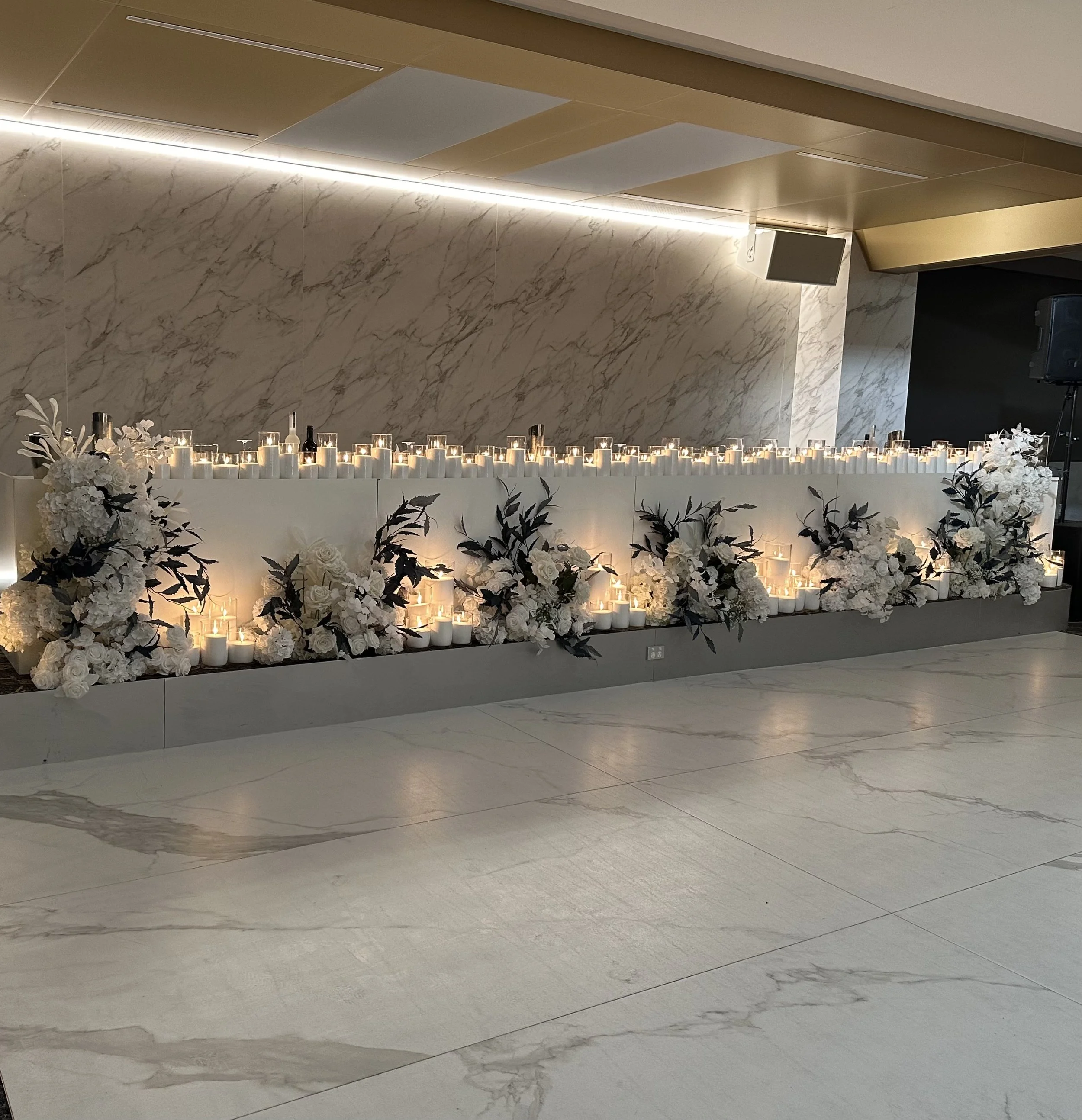 Decorative indoor setup with a table adorned with white candles and white flowers, set against a marble wall with warm lighting and a black speaker in the background.