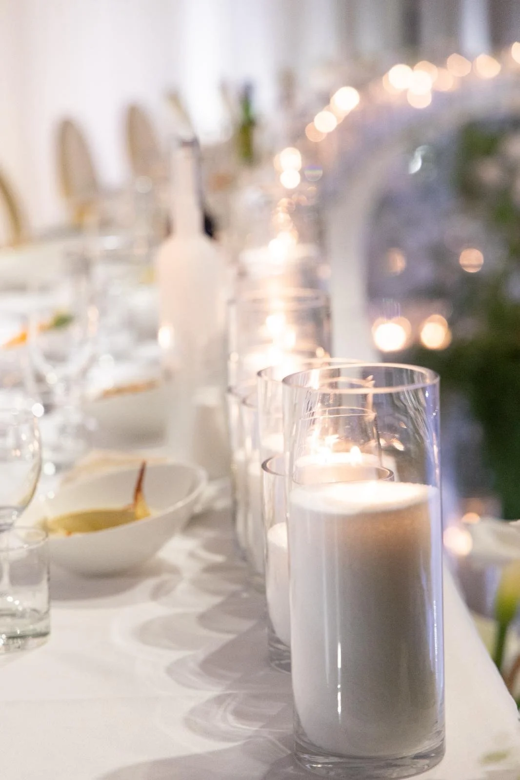 A close-up of white candles in tall glass holders on a dining table, with a blurred background of more candles and tableware, creating a cozy and elegant ambiance.