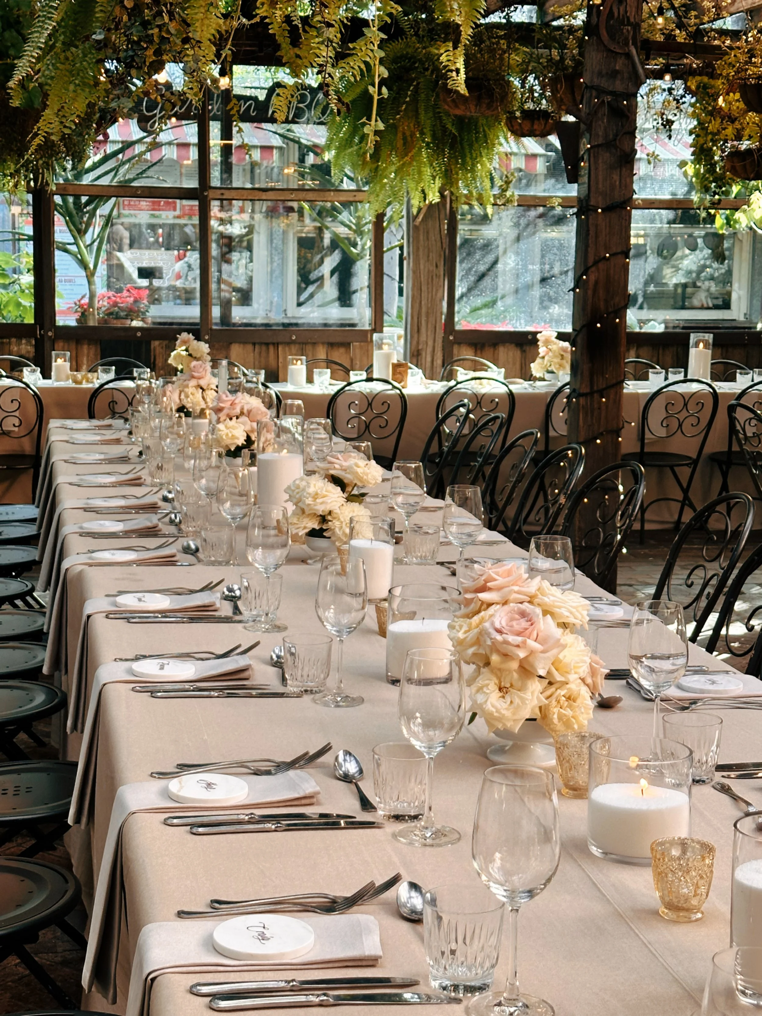 A banquet-style table decorated with pink and white flowers, candles, and glassware, set for a formal event or wedding reception, in a rustic venue with wooden beams and greenery.