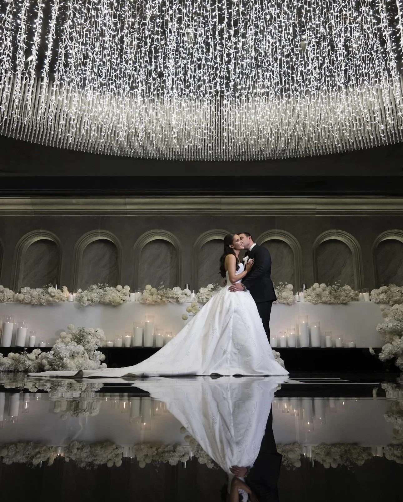 A bride and groom sharing a kiss in a wedding reception hall decorated with white flowers and candles, a large chandelier above them, and a dark wall with arched panels in the background.