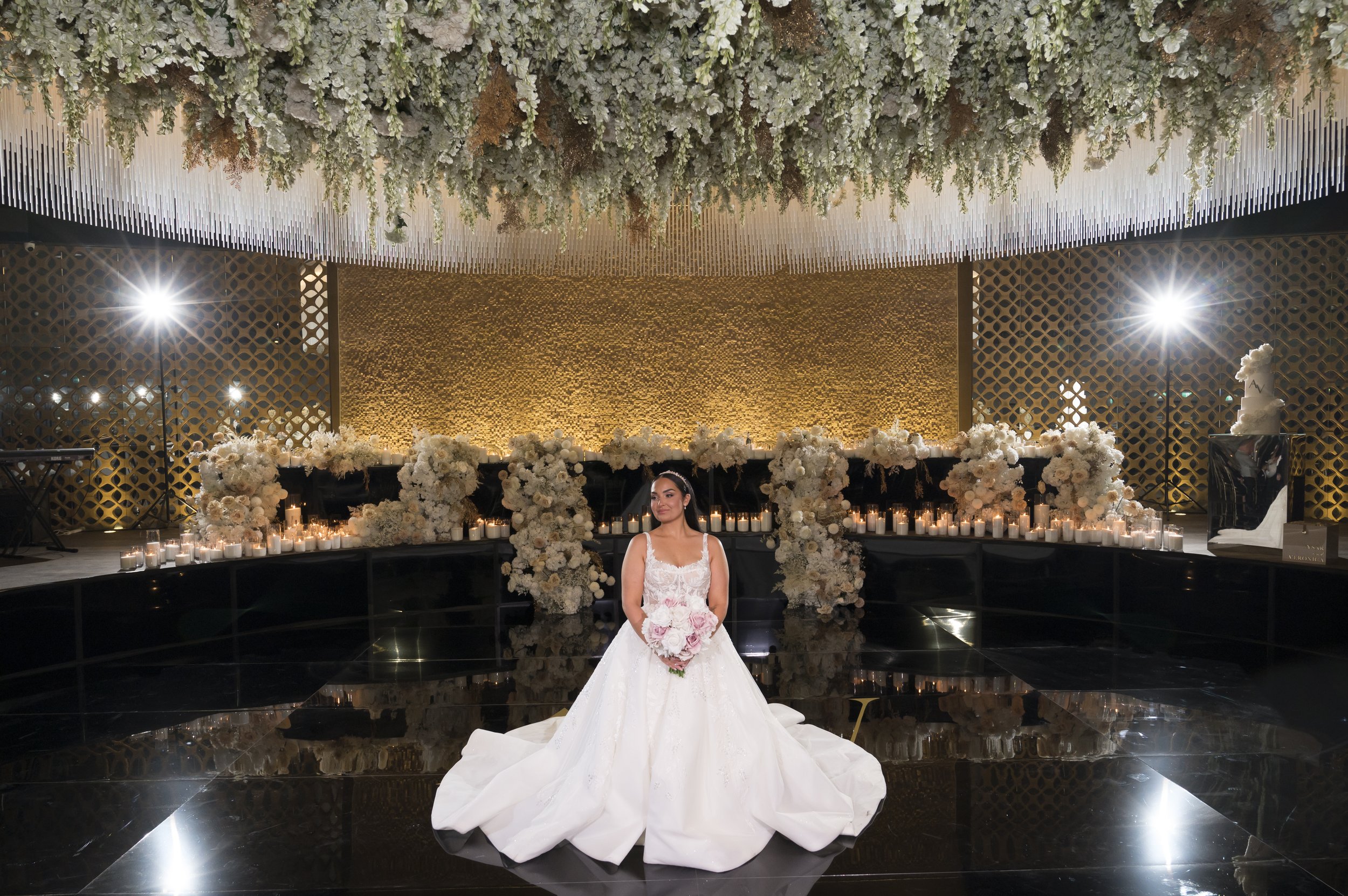A bride in a white wedding gown holding a bouquet of pink roses poses in front of a decorated wedding reception area with white flowers and candles, in a venue with a gold and black color theme and floral ceiling decor.