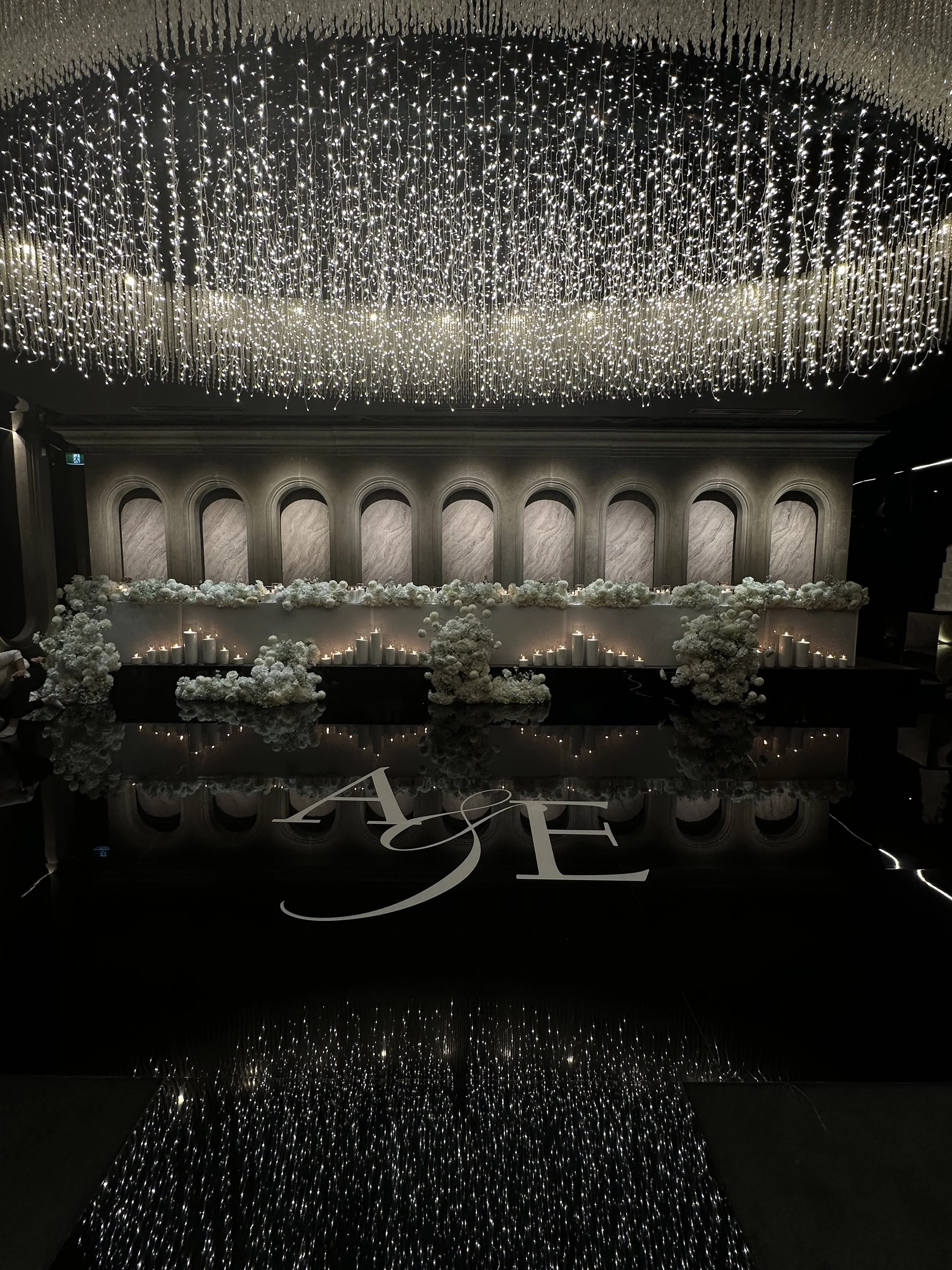 A grand indoor setting decorated with a large, intricate chandelier of hanging fairy lights from the ceiling. The lower part of the image features an arrangement of white floral decorations and candles on a long table. The background has a textured w