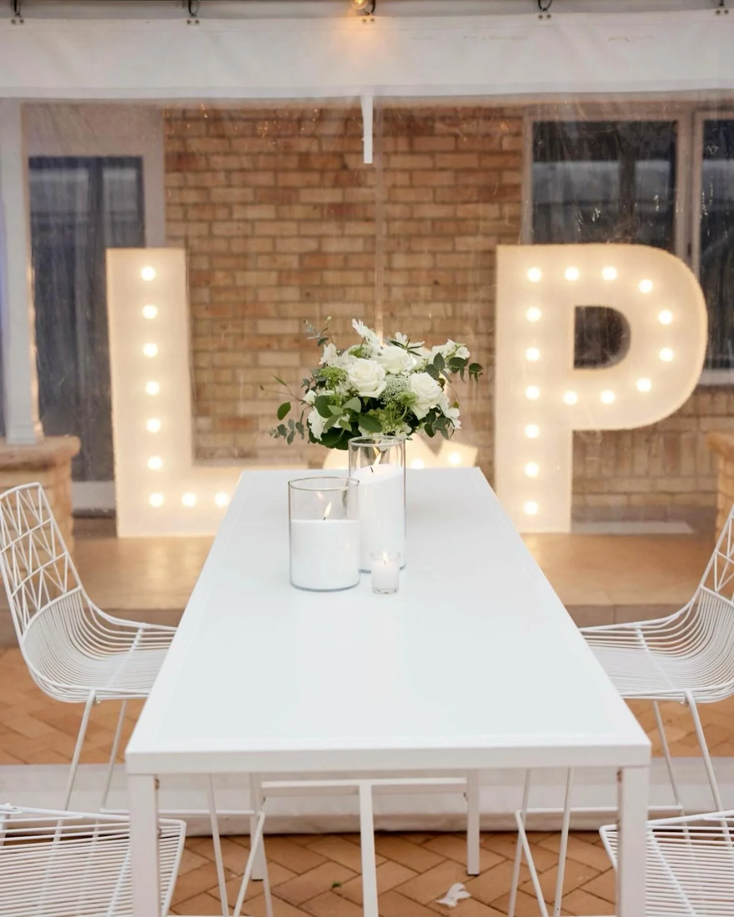 A decorated event setup with a rectangular white table, floral centerpiece, and large illuminated letter 'L' and 'P' in the background, against a brick wall with windows.