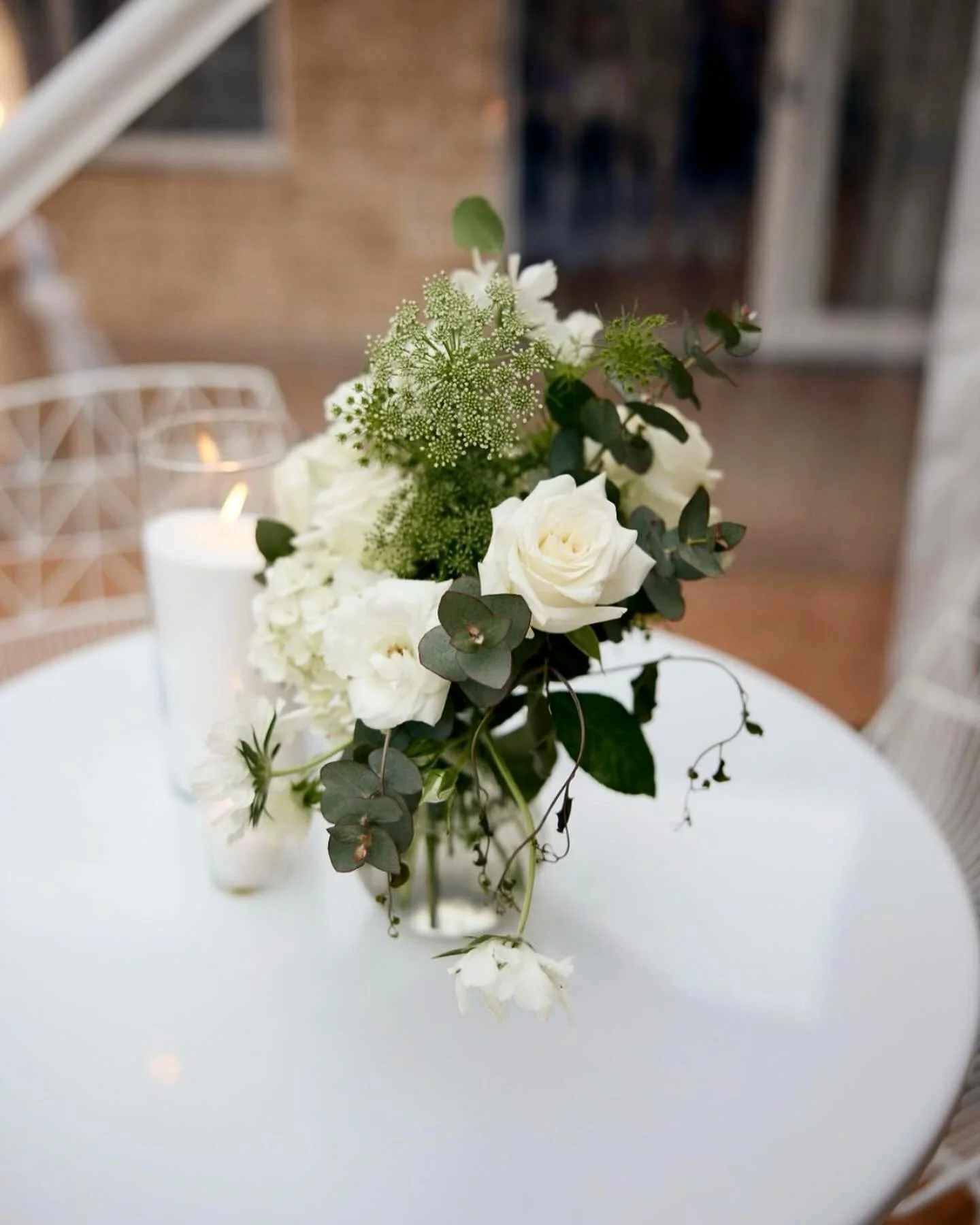 A floral centerpiece on a white table with white roses, green foliage, and white flowers, with a candle in a glass holder in the background.