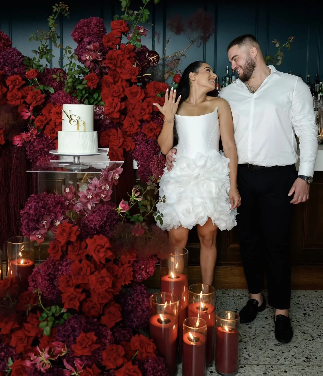 A couple dressed in wedding attire standing in front of a floral arrangement with a cake and sand candles, smiling at each other.