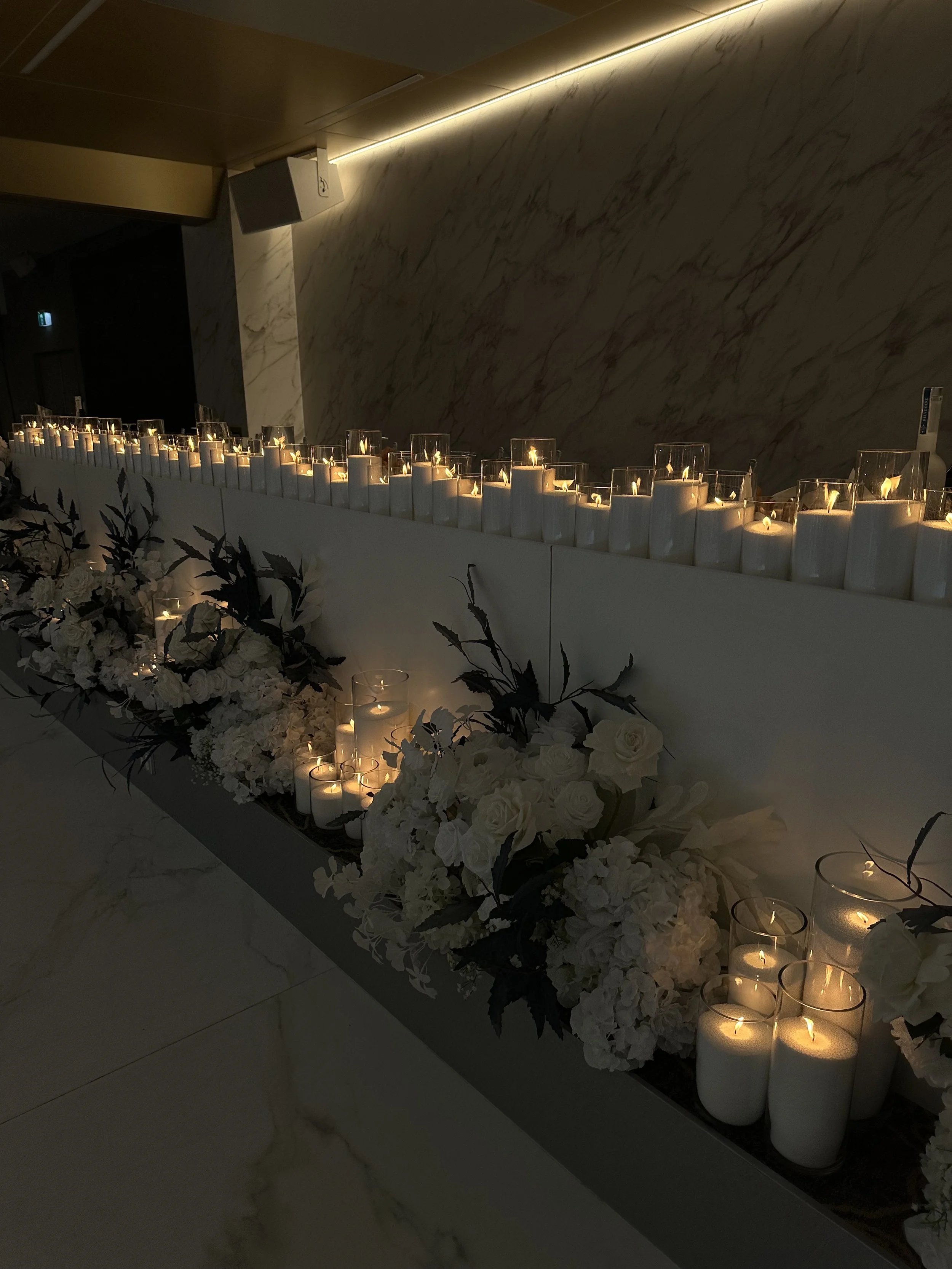 Multiple white candles in glass holders and larger white pillar candles arranged on a dark surface with white flowers and black foliage in front of a marble wall, creating a decorative display.