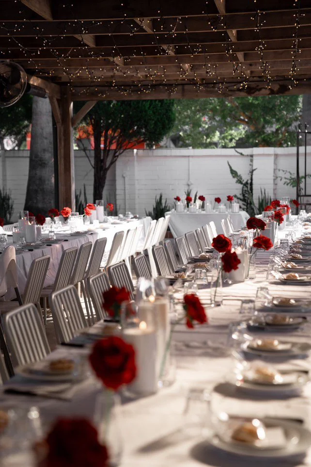 Long banquet table set outdoors with white tablecloths, red roses in vases, white sand candles, plates, glasses, and napkins, under a wooden pergola decorated with string lights.