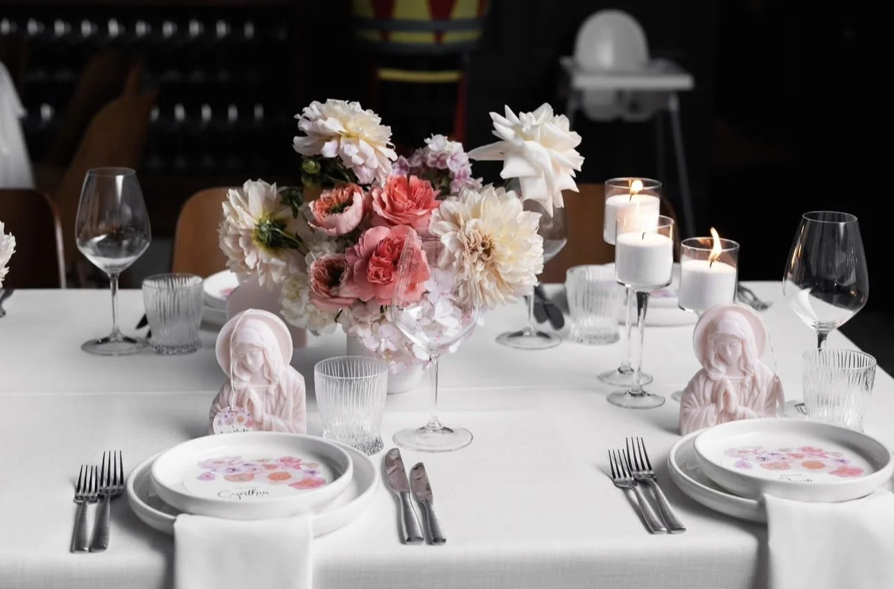 Elegant table setting with floral centerpiece, pink and white flowers, lit candles, tea light holders, pink figurines, floral-patterned plates, white napkins, and glassware.