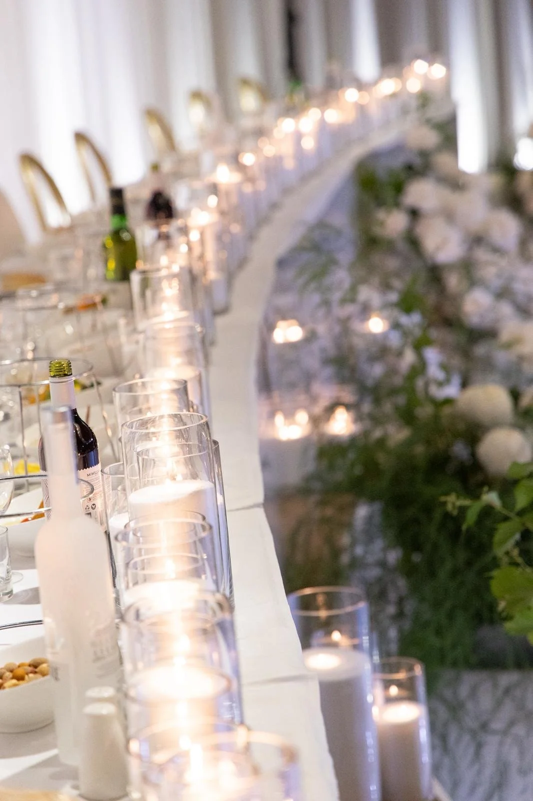 A long banquet table decorated with candles, glassware, and floral arrangements, with chairs on one side and a wall of white curtains in the background.