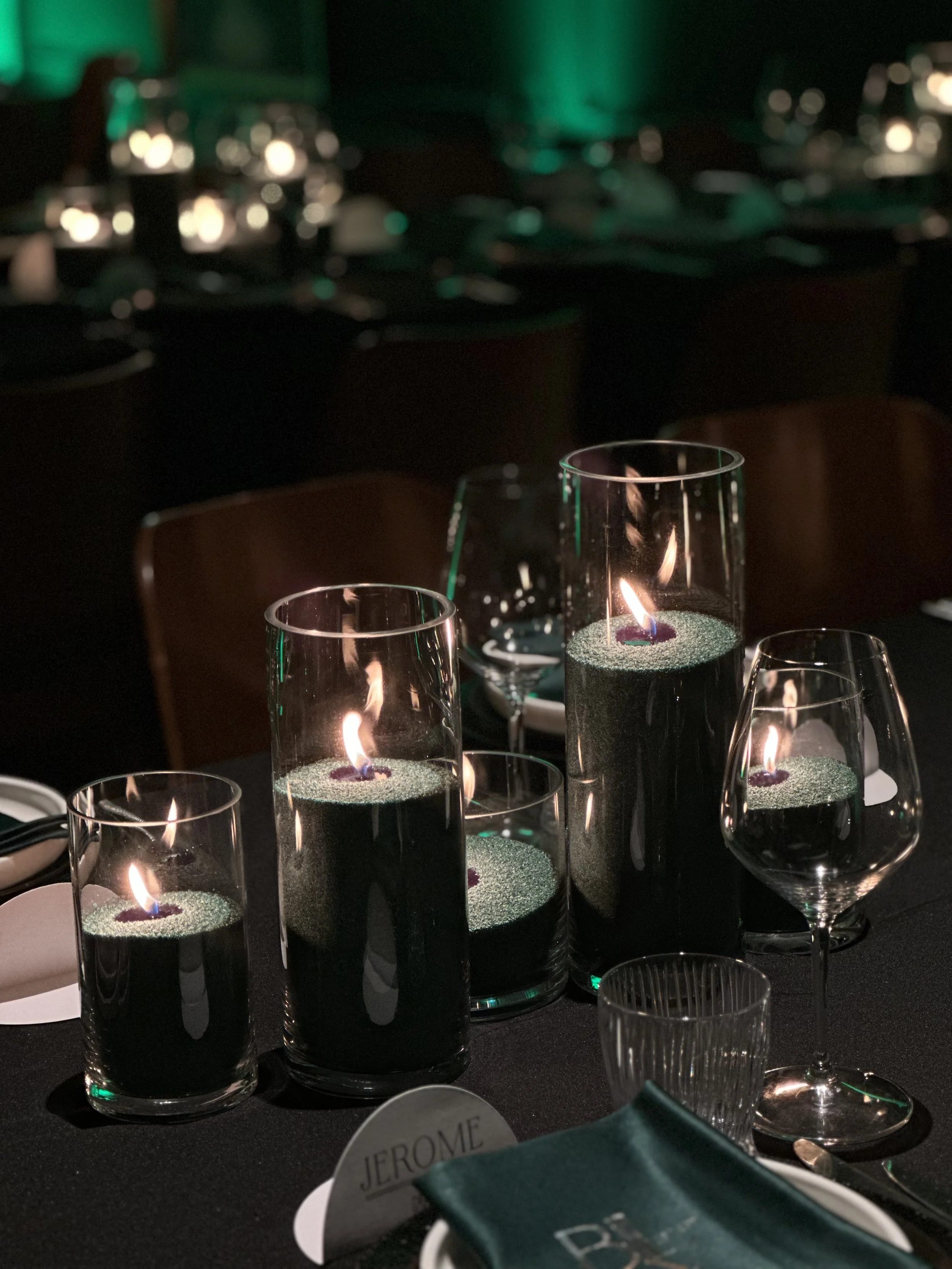 Several lit candles in glass holders on a table at night, with wine glasses around and a dark background.