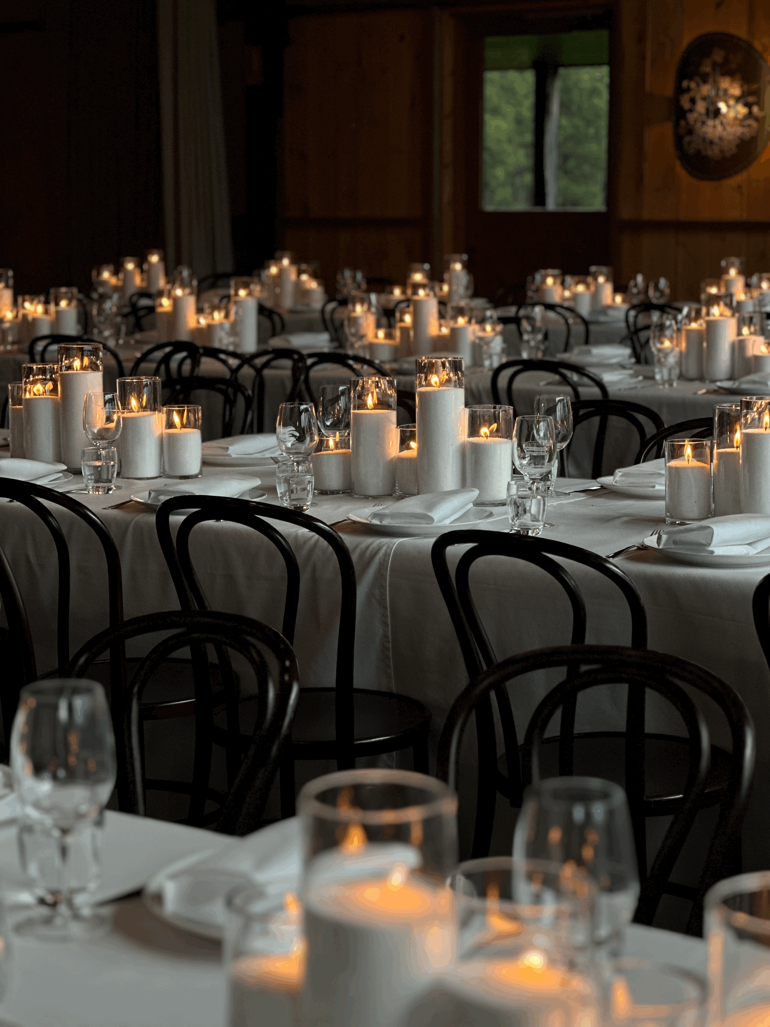 A dimly lit banquet hall with long dining tables decorated with tall candles, wine glasses, and white napkins. The walls are made of wood, with one window showing greenery outside. There are black chairs around the tables, creating an elegant atmosph