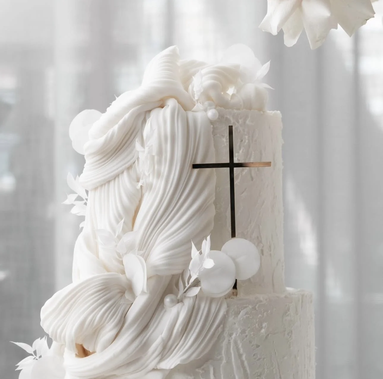 White religious-themed cake with ruffled frosting, a black cross on the side, and floral decorations.