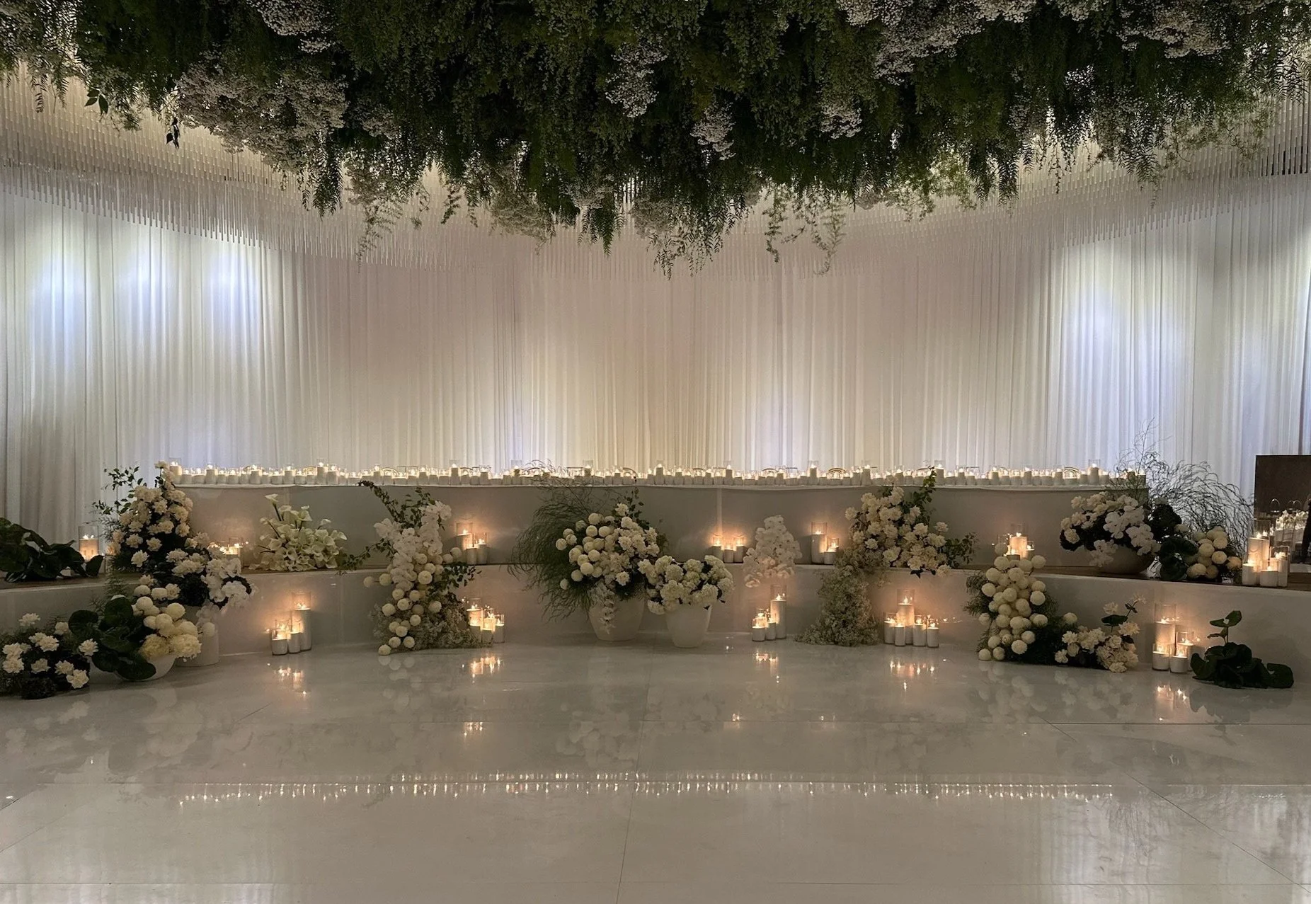 Elegant wedding ceremony backdrop decorated with white flowers, white sand candles, and draped white curtains.