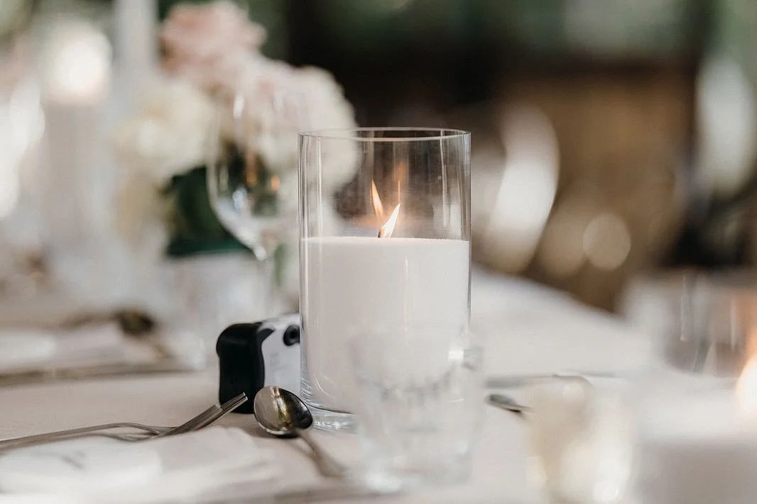 A lit candle inside a clear glass on a dining table with silverware and blurred background.
