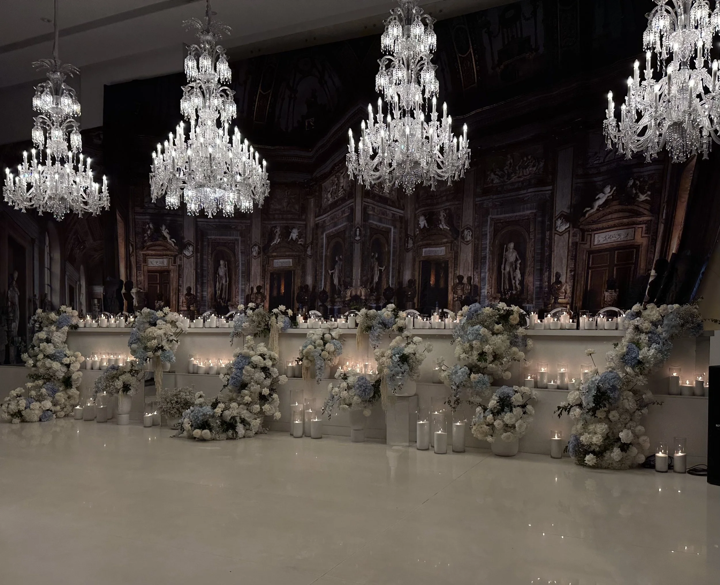 Elegant event space decorated with tall crystal chandeliers hanging from the ceiling, and floral arrangements and candles on a stage or table in front, with a dark ornate wall in the background.