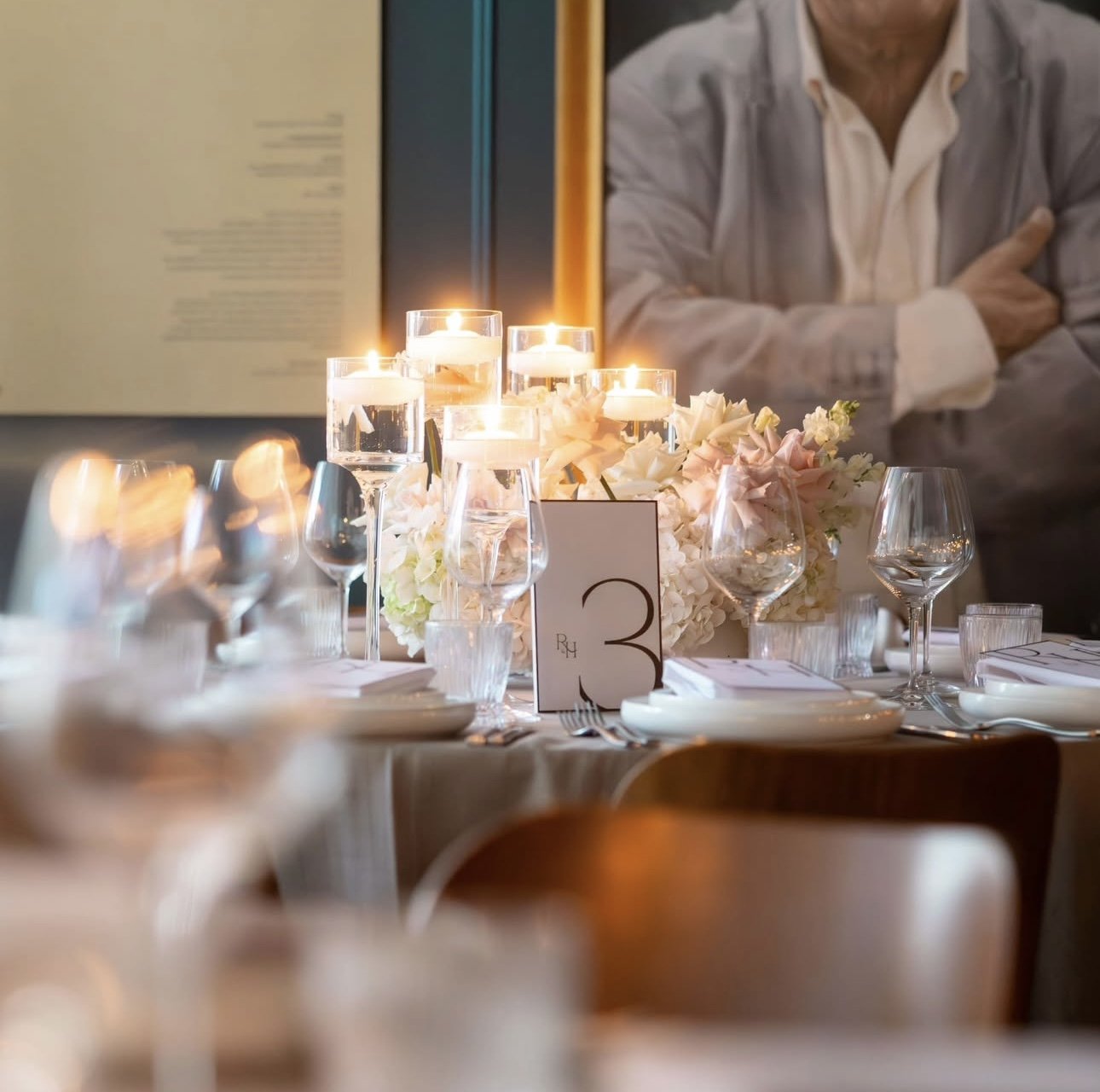 A beautifully set elegant dinner table with lit candles, glassware, floral arrangements, and a table number card, in a formal event setting.