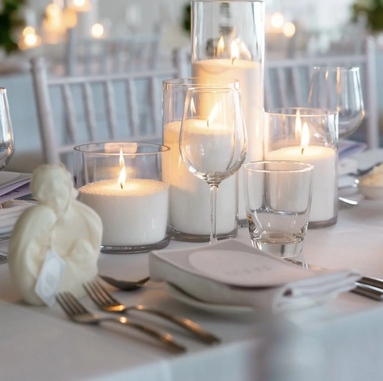 A table decorated with white candles in glass holders, empty wine glasses, a white napkin, and a white carved flower, set for a formal dining event.