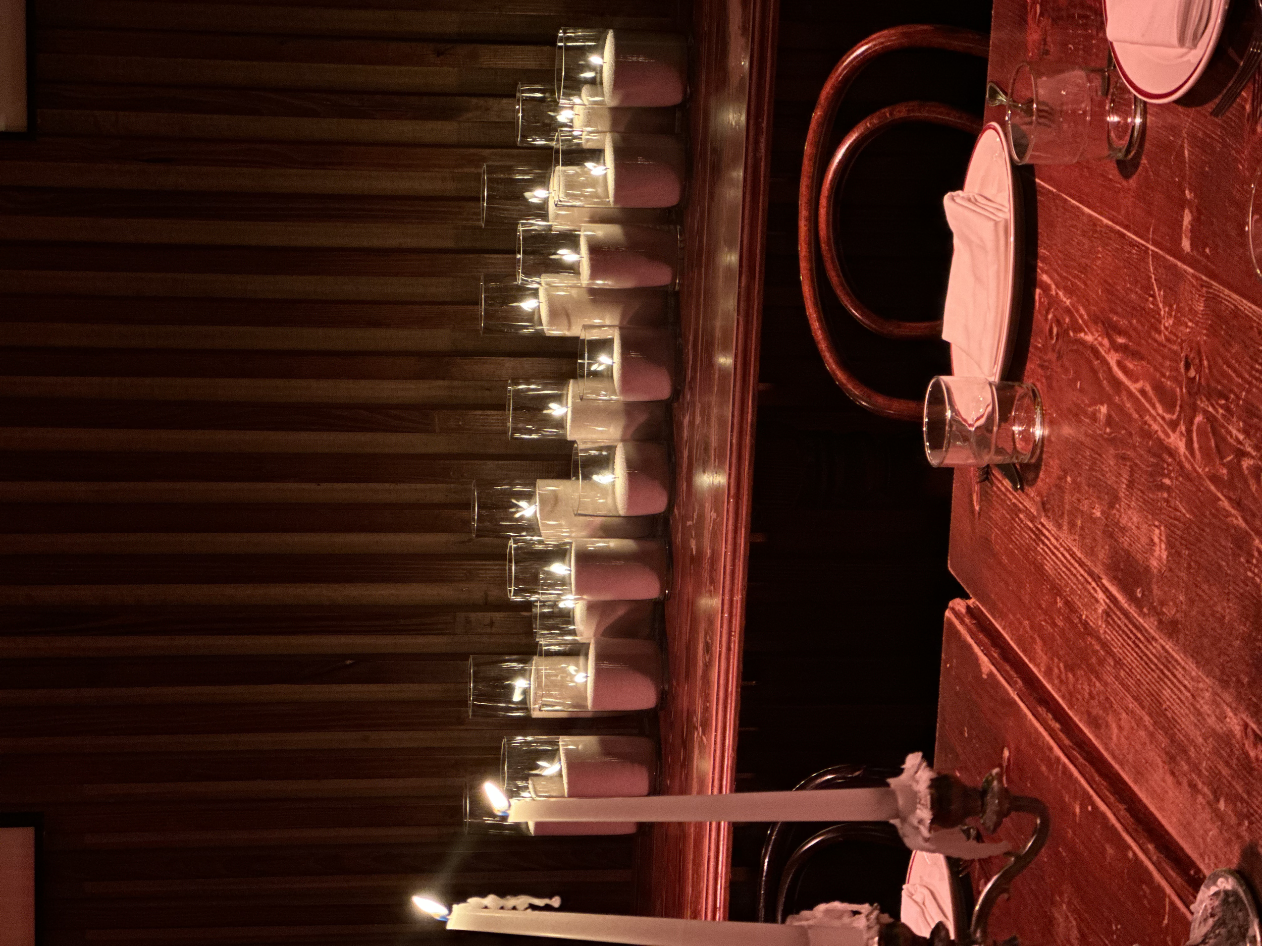 A dimly lit restaurant or dining room with a wooden table set with glasses and plates, and candles as part of the table decor. Vertical wooden paneling is visible on the wall in the background.