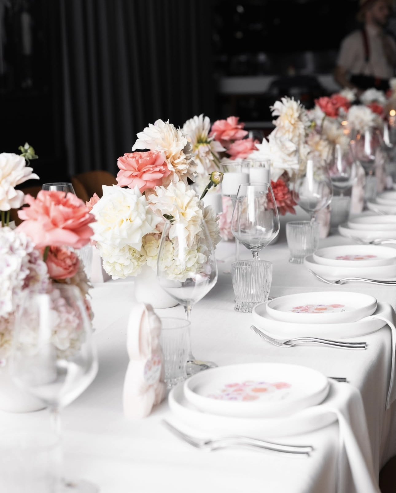 Elegant table setting with white tablecloth, floral centerpieces in vases, wine glasses, plates with pink flower designs, and silverware for a formal event.