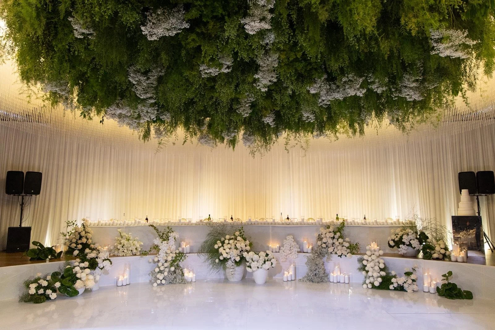 An elegant indoor event venue decorated with white flowers and candles, featuring a long table with floral arrangements and candles, and a large floral centerpiece hanging from the ceiling.
