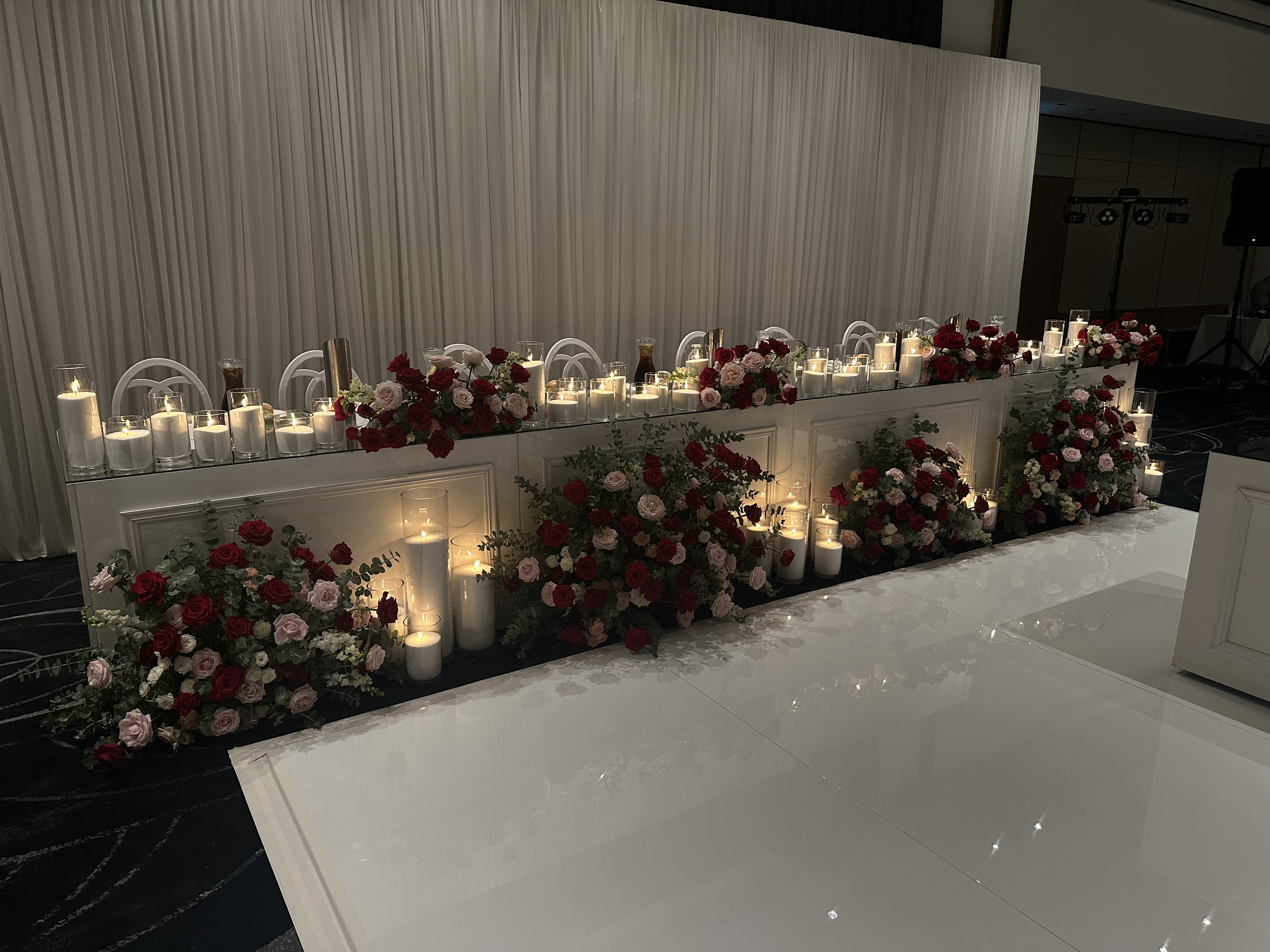 Elegant event setup with a long white table decorated with red and pink roses, surrounded by candles in glass holders, and a plain cream-colored curtain backdrop.