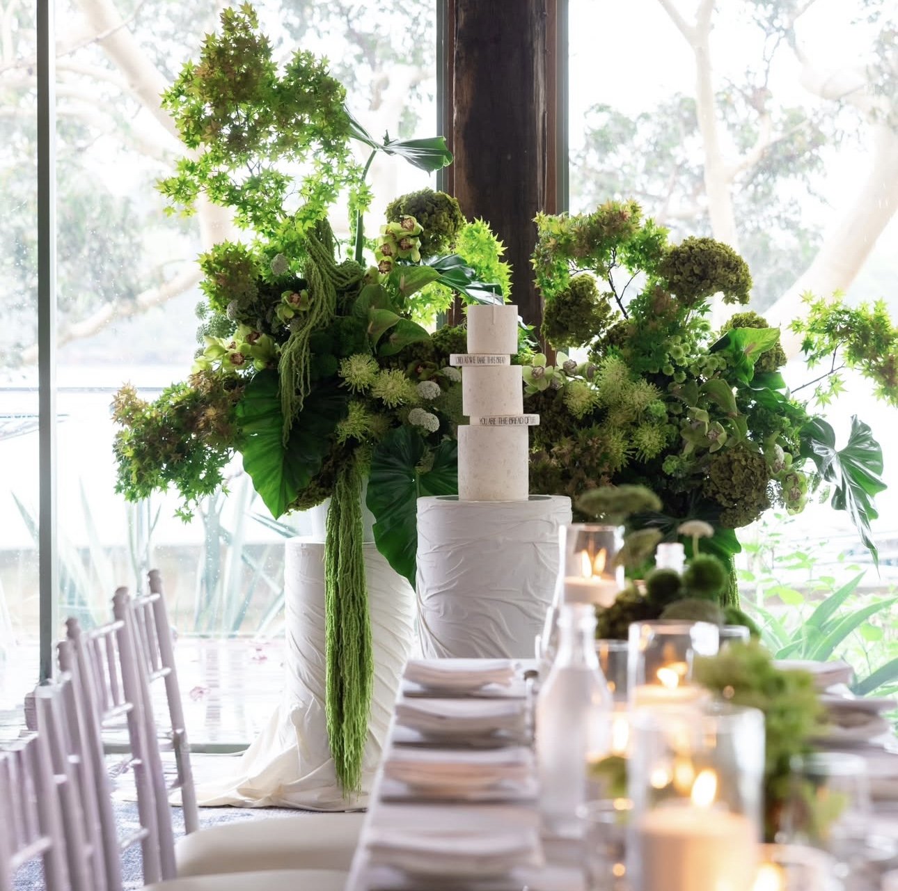 Elegant table setting with large floral arrangements in white vases, lit candles, and neatly stacked plates against a large window with trees outside.