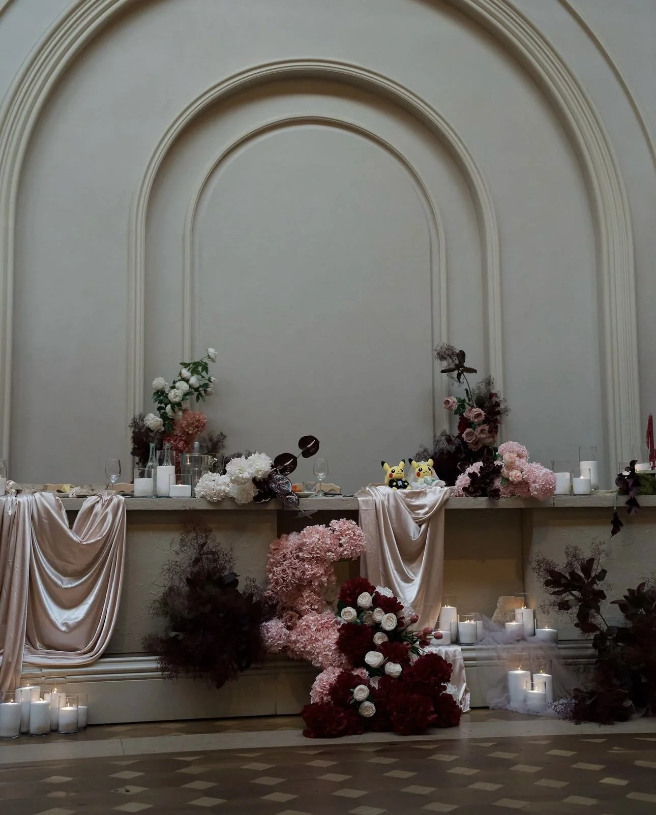 Decorative floral display on a table set against a beige wall with arched moldings, including pink, white, and burgundy flowers, candles, and plush Pikachu toys, with draped fabric and lit candles on the floor.
