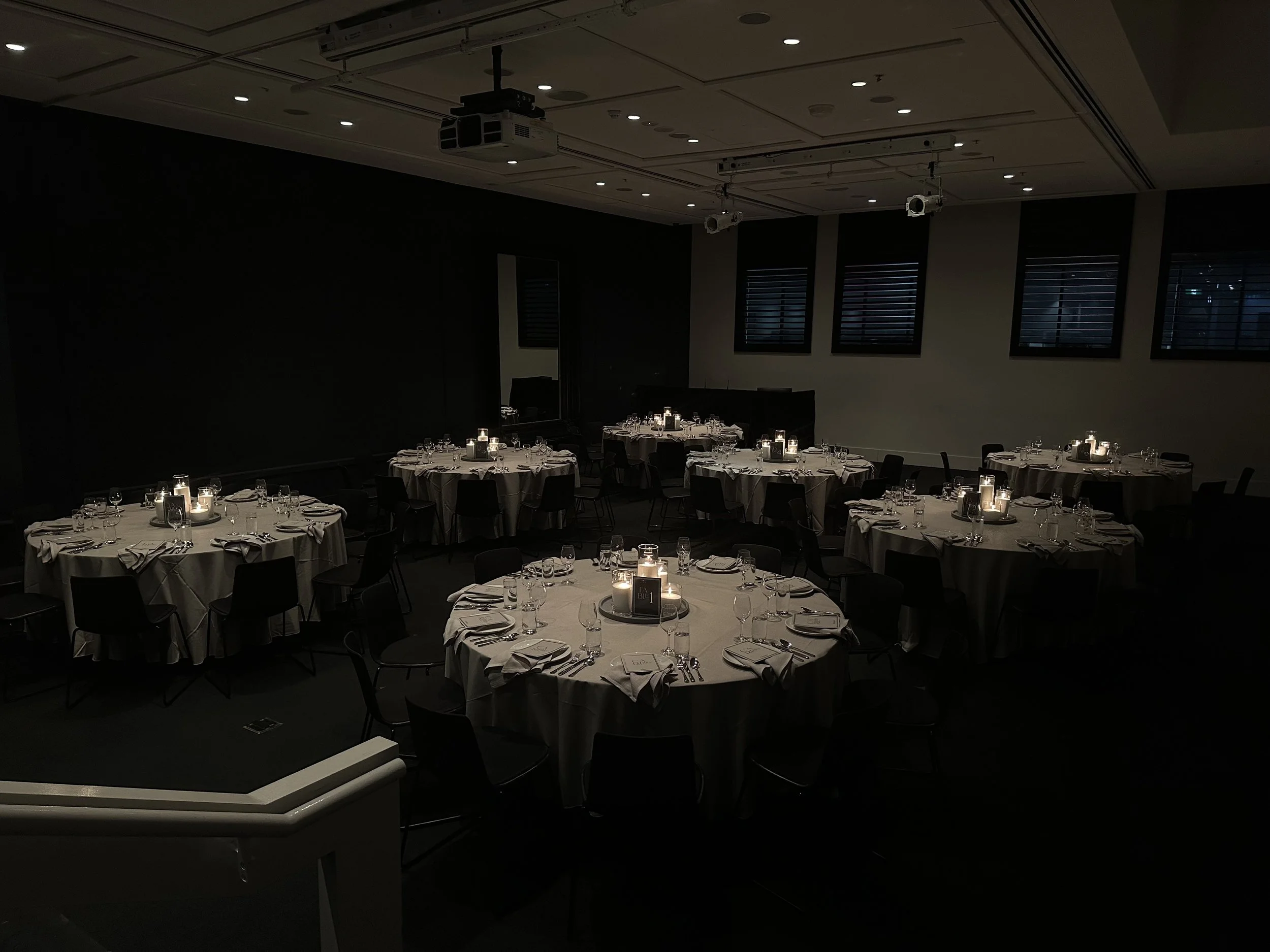 A dimly lit banquet hall with circular tables draped in white tablecloths, set with glassware and plates, and illuminated by small candles.