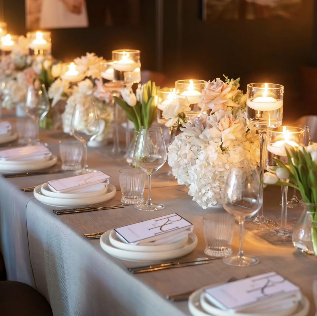 Elegant dining table set with white plates, silverware, wine glasses, decorated with white floral arrangements and floating candles in glass holders, in a warm, inviting atmosphere.
