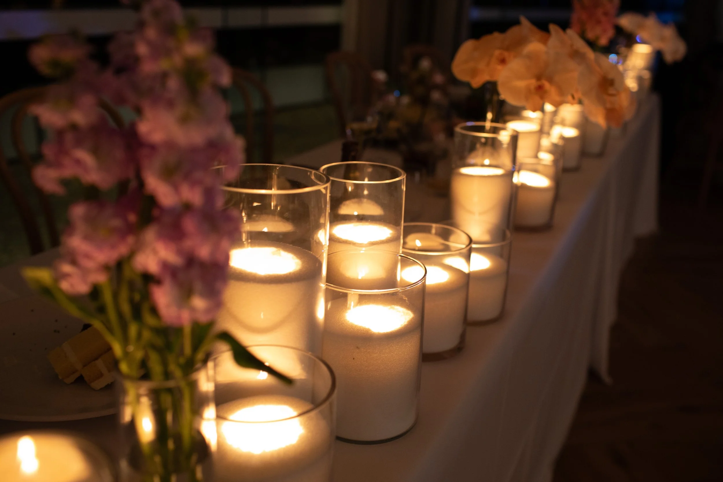A lineup of white candles in glass holders providing soft candlelight, with pink and peach flowers in the foreground, likely part of a wedding or romantic event decor.