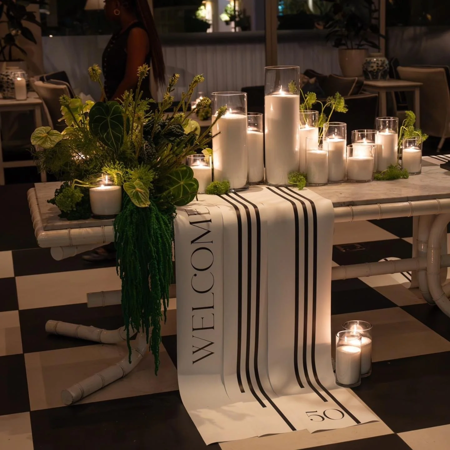 A white bamboo table decorated with white candles in glass holders and a green floral arrangement, with a white and black striped "Welcome" runner.