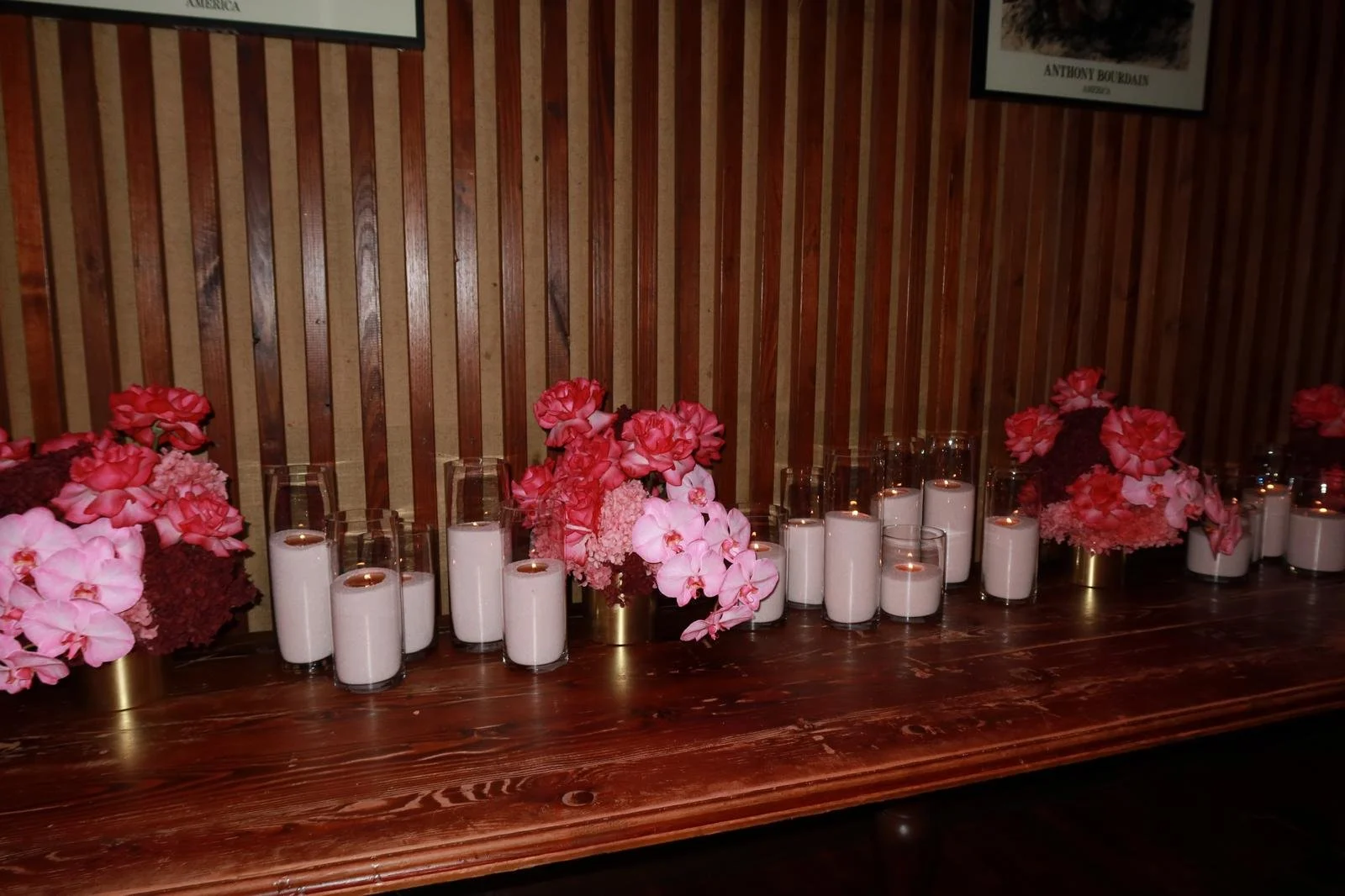 Flowers in pink and red shades with white candles in glass holders on a wooden table against a wooden paneled wall.