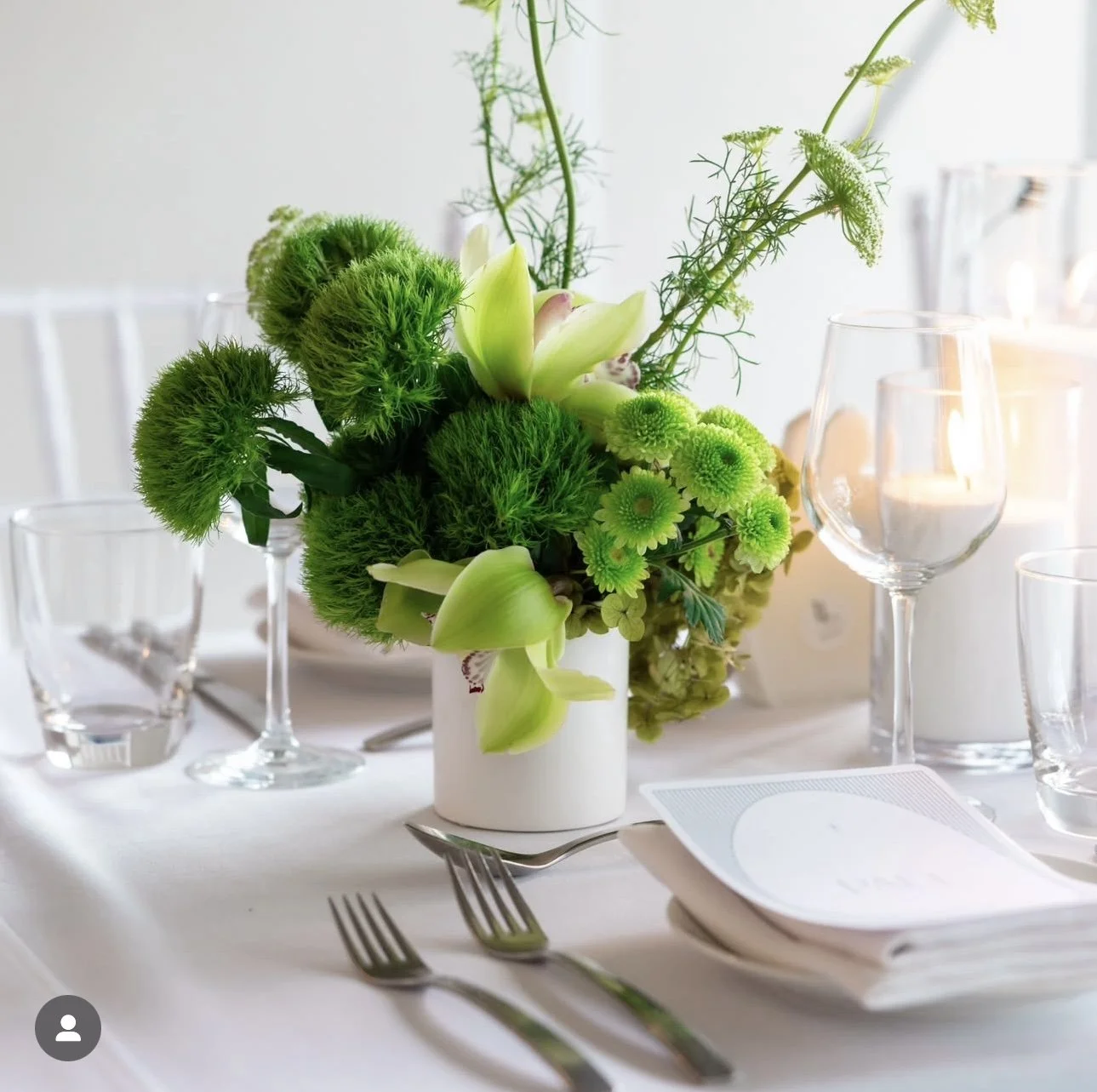 Elegant table setting with a white vase floral centerpiece featuring green flowers, surrounded by wine glasses, water glasses, silverware, and a napkin with a menu on a white tablecloth.