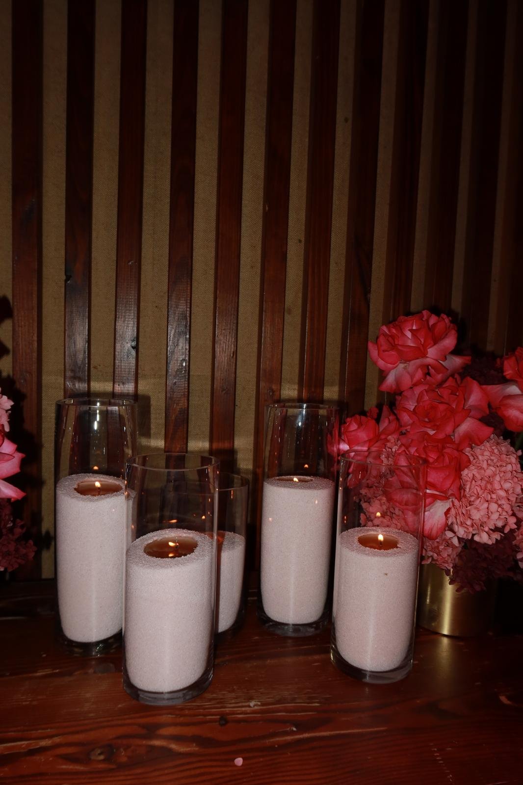 Four tall glass candle holders with white candles and flickering flames, pink floral arrangements in gold vases, and a wooden slat background.