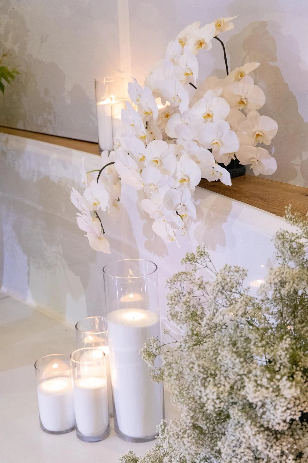 White orchids and baby's breath flowers on a table with candles in glass holders and a lit candle in a tall glass