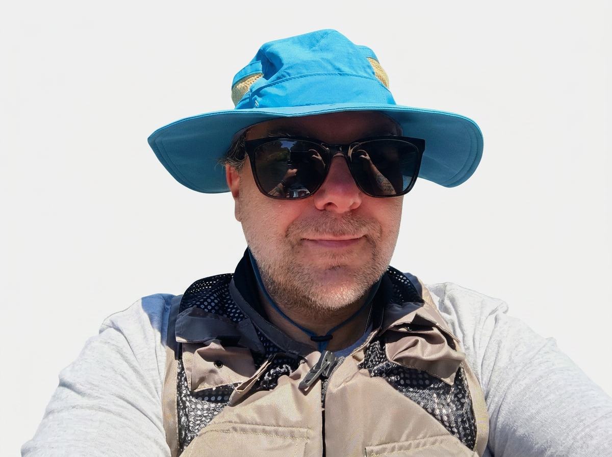 A man wearing a large blue sunhat, black sunglasses, a gray shirt, and a beige and black hiking vest.