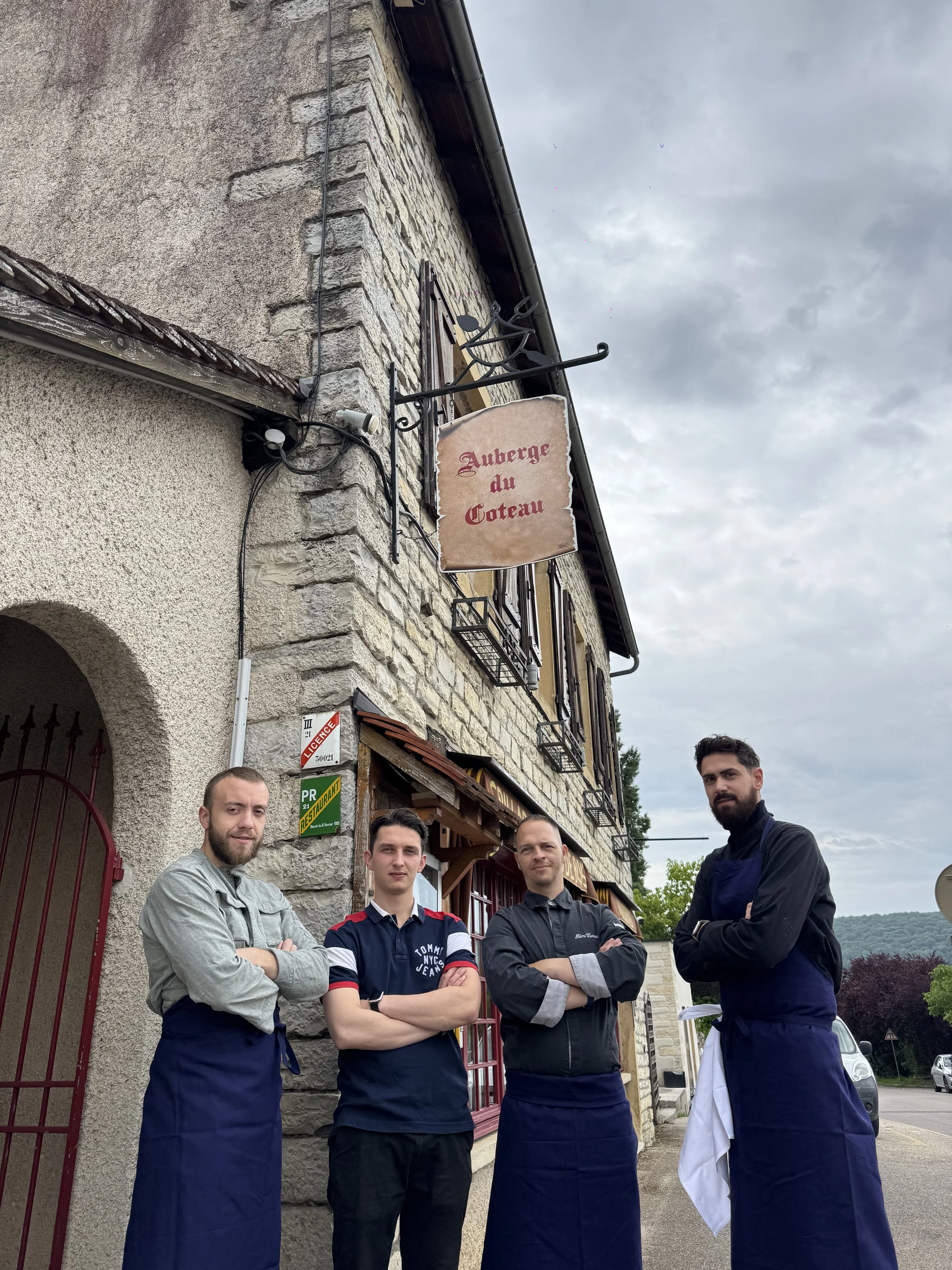 Quatre hommes debout devant un restaurant en pierre, deux portent des tabliers bleus, un porte un polo, un porte une chemise et un tablier gris, tous avec les bras croisés, devant un bâtiment avec une plaque signalisant un établissement certifié.