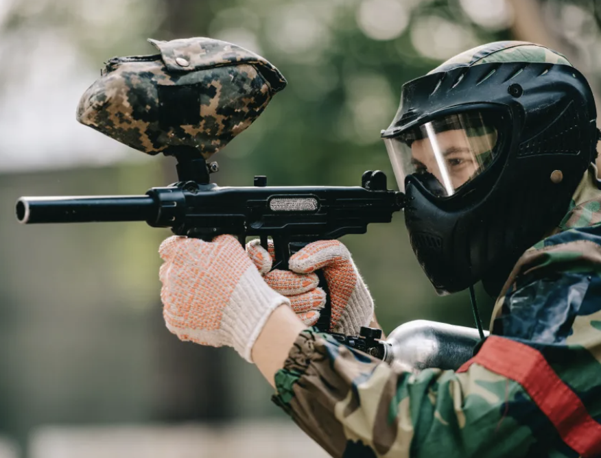 Un soldat en tenue camouflage, portant un casque et un masque facial, tenant une arme à feu avec une cible camouflée attachée à l'arme, dans un environnement forestier.