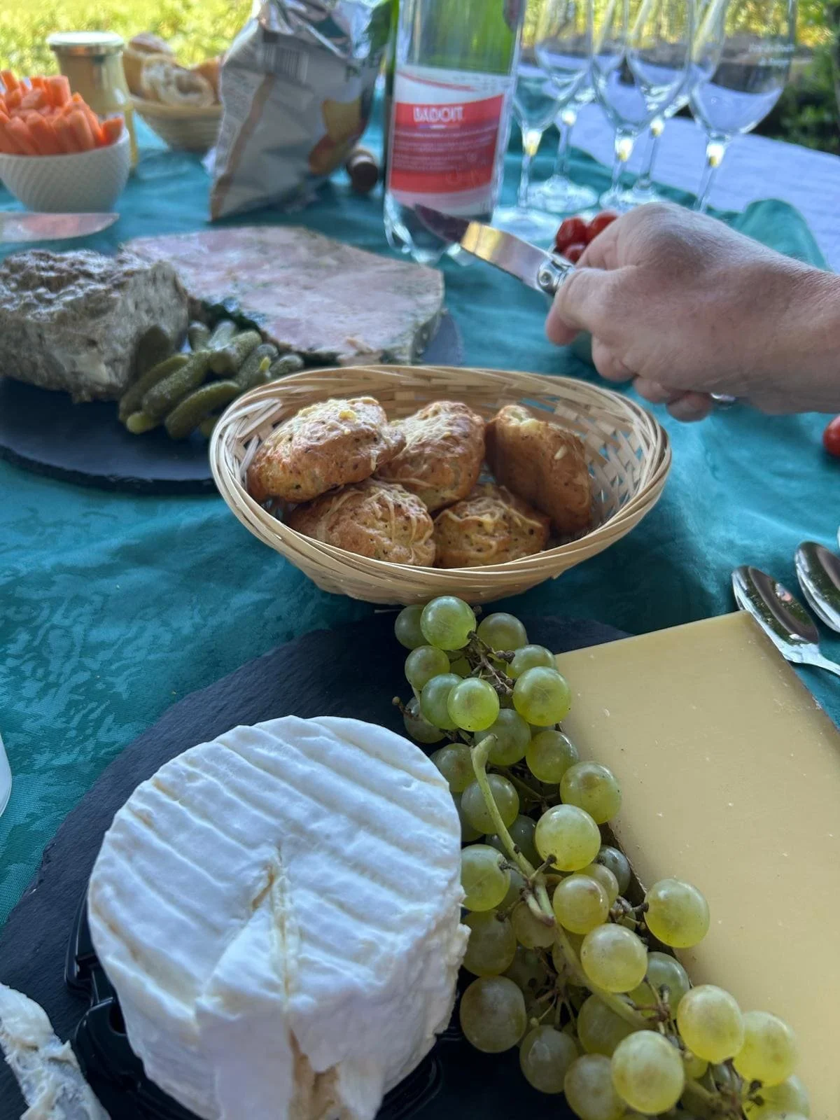 Un plateau de fromages avec un brie, des raisins verts, et du fromage jaune, une table avec des platés de charcuterie, des petits pains, et des snacks, lors d'un repas en extérieur.