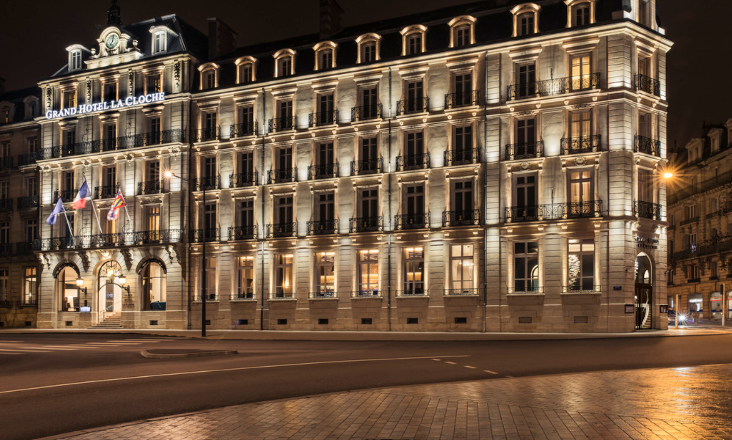 Façade illuminée de l'hôtel Grand Hôtel La Cloche à Troyes la nuit avec drapeaux français, européen et régional à l'entrée.