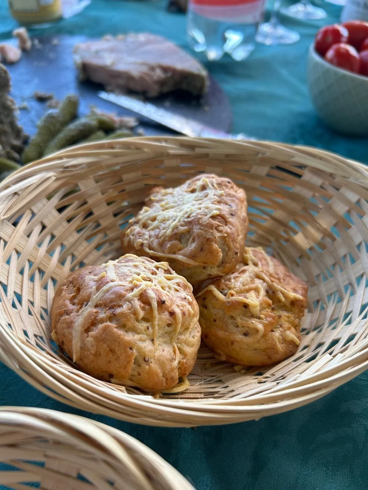Trois muffins dans un panier en osier, garnis de fromage, avec un fond flou montrant de la viande, des tomates cerises, un verre d'eau, et une table avec un tissu bleu.