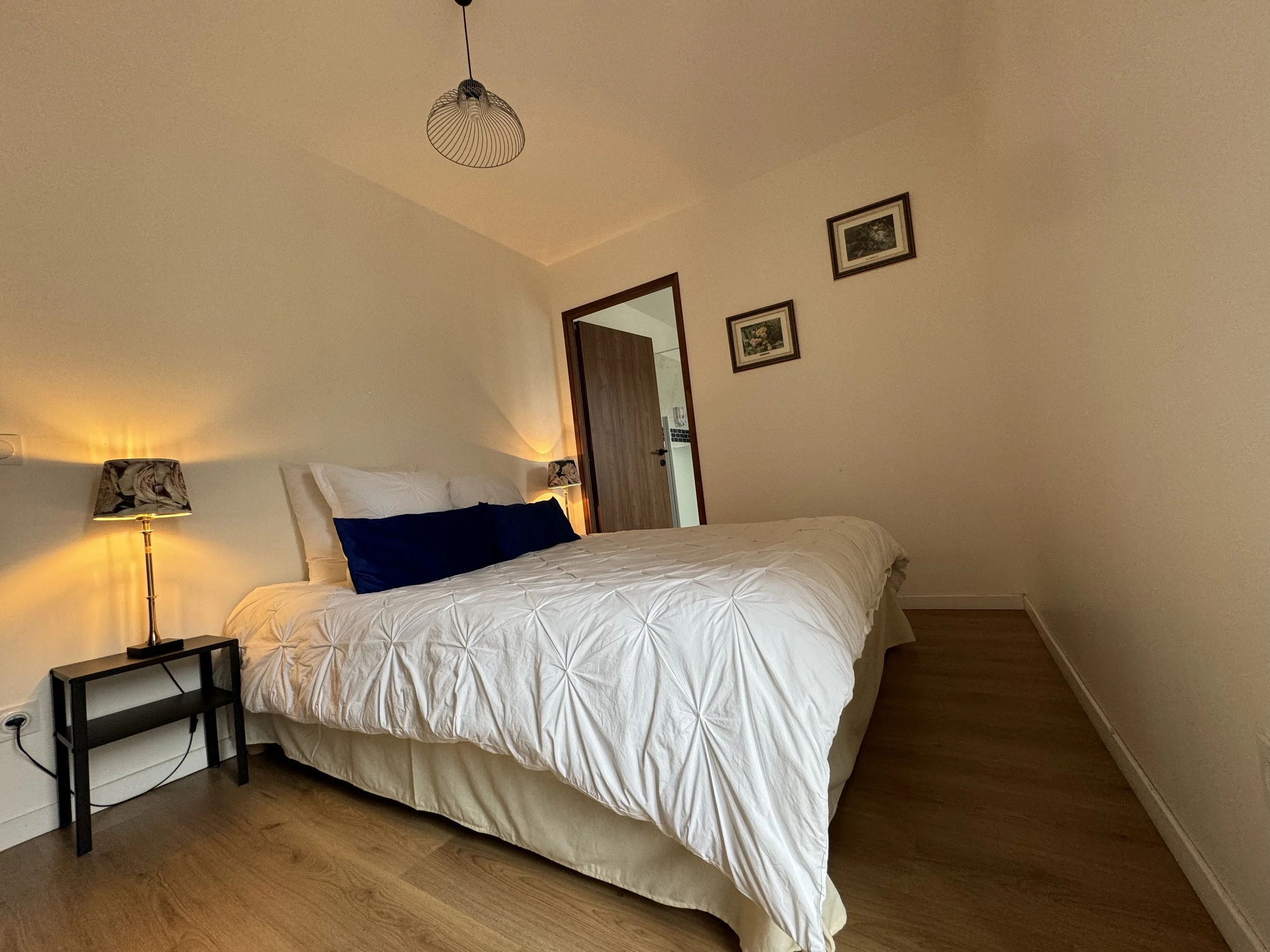 Chambre à coucher avec lit double, draps blancs, deux lampes de chevet allumées, une décoration avec trois cadres, et une porte en bois.
