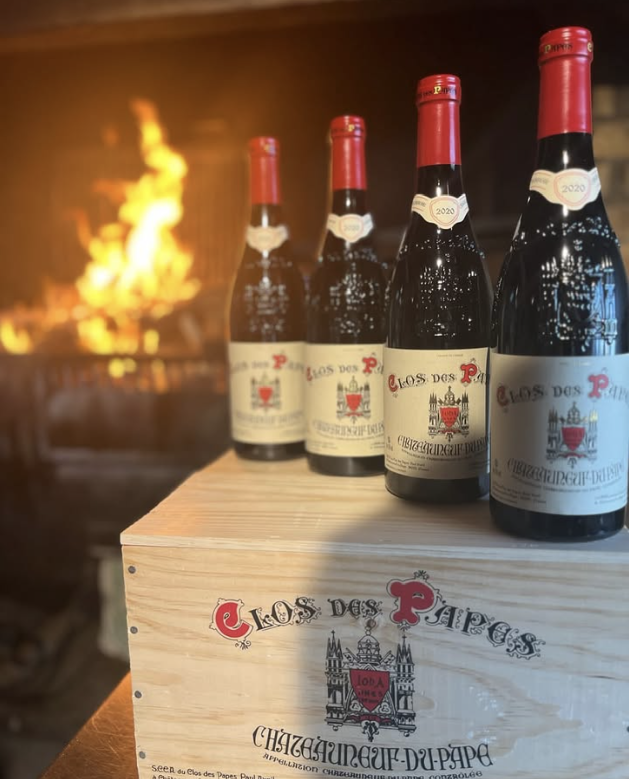 Quatre bouteilles de vin rouge sur une caisse en bois portant l'inscription 'Clos des Papes', avec un feu de cheminée en arrière-plan.