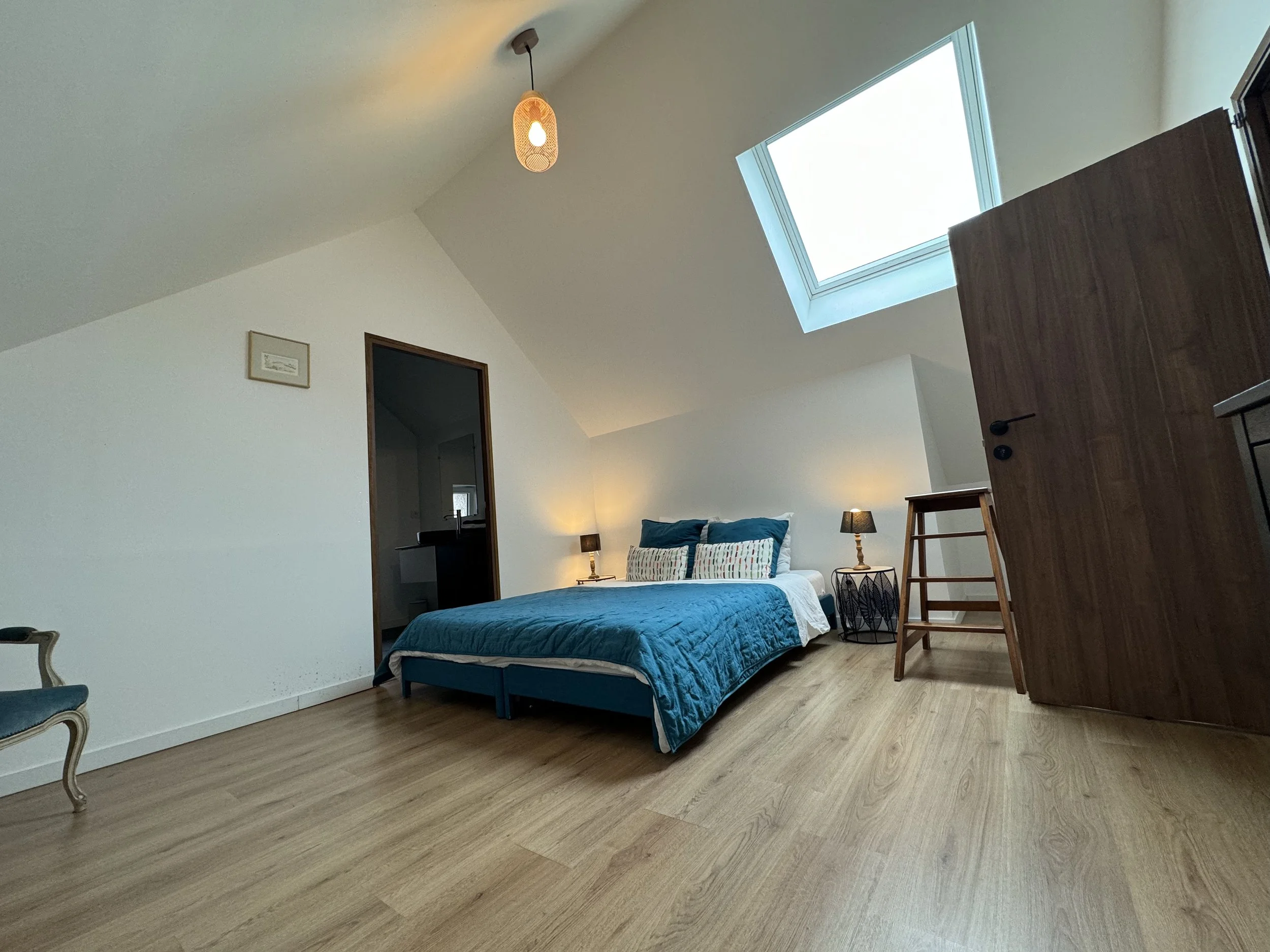 Chambre moderne avec lit double, lampe de chevet, plafond avec fenêtre de toit, meubles en bois et sol en bois clair.