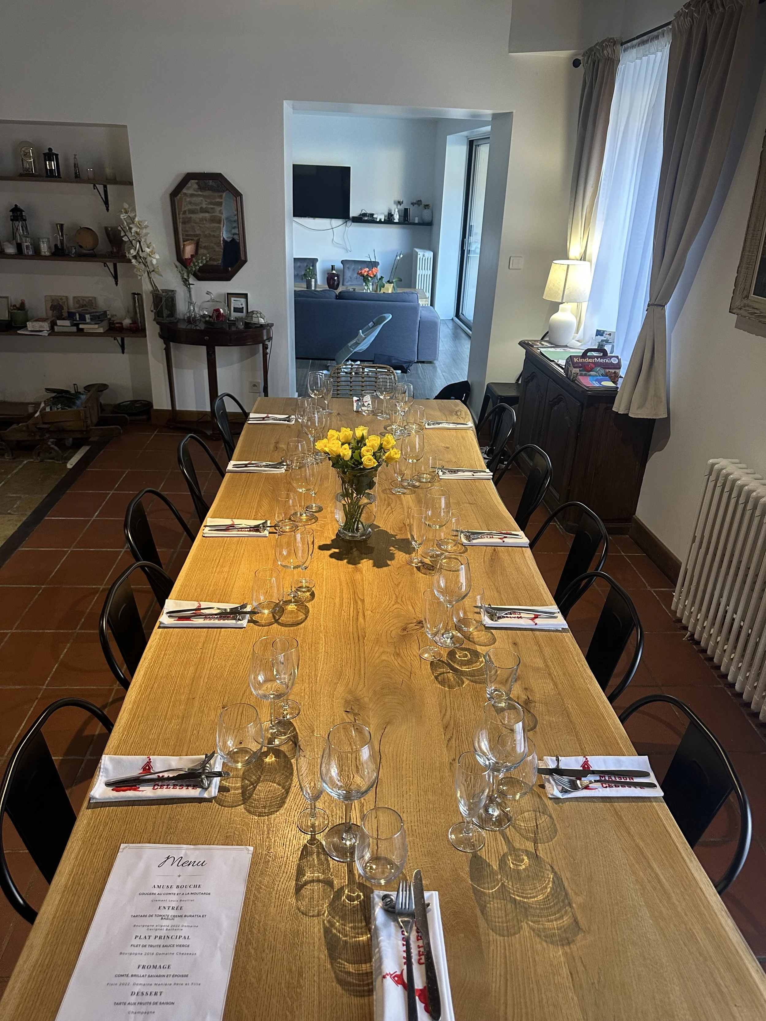 Une grande table en bois avec des verres, des assiettes, des serviettes, et un bouquet de fleurs jaunes au centre, prête pour un repas. La pièce a des murs blancs, avec une cheminée et des étagères décoratives à gauche, et abrite un salon visible à t