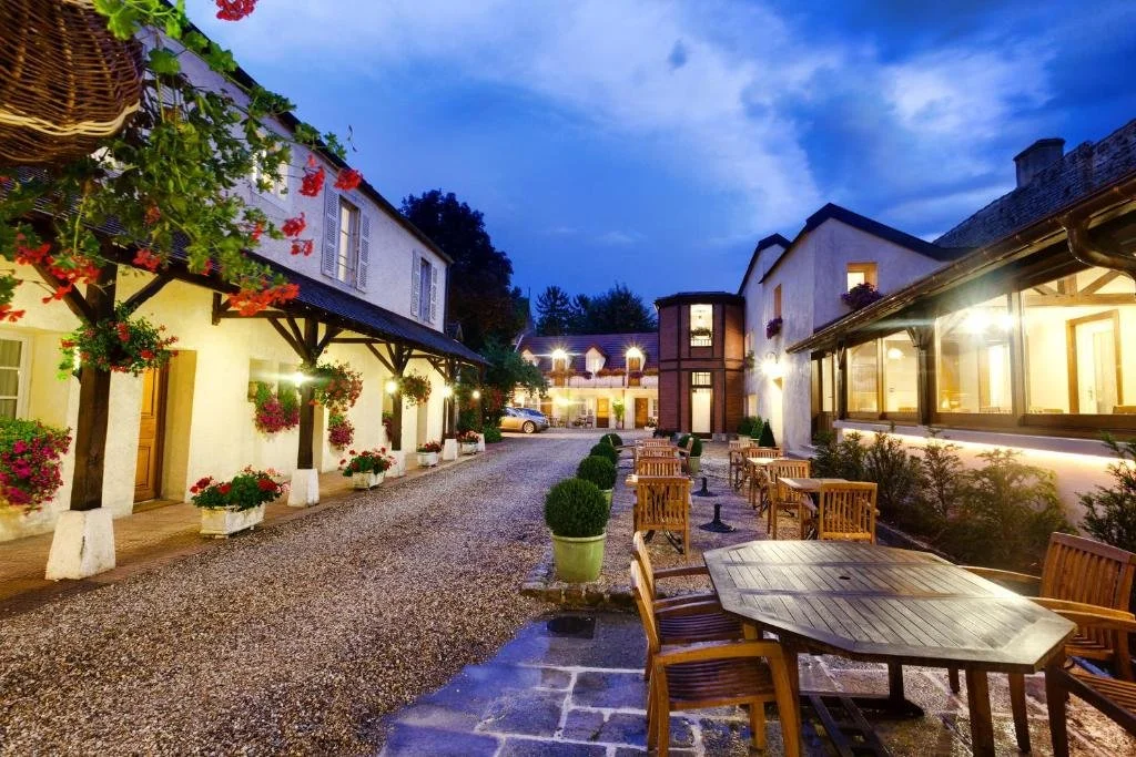 Une cour extérieure éclairée avec des tables en bois et des chaises, entourée de bâtiments blancs avec fenêtres, décorée de fleurs et de plantes