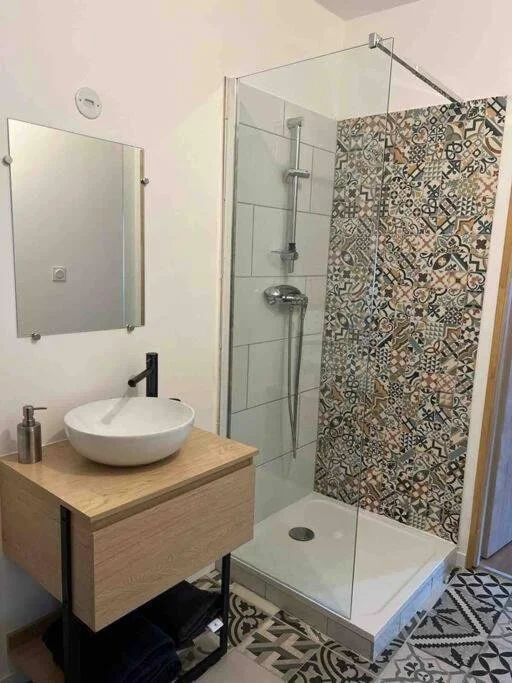 Salle de bain avec une cabine de douche en verre, un lavabo sur un meuble en bois, un miroir mural, un porte-savon, et un sol avec des carreaux de motifs géométriques.