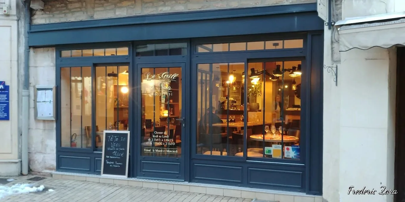 Vitrine d'un restaurant ou café avec une façade en métal bleu foncé. À l'intérieur, des tables sont dressées, quelques verres à vin sur une table et une ambiance chaleureuse avec un éclairage doux. Devant, une ardoise indique une offre spéciale de soupe de veau à 8 euros. Il fait probablement froid dehors, avec un petit tas de neige à gauche sur le trottoir.