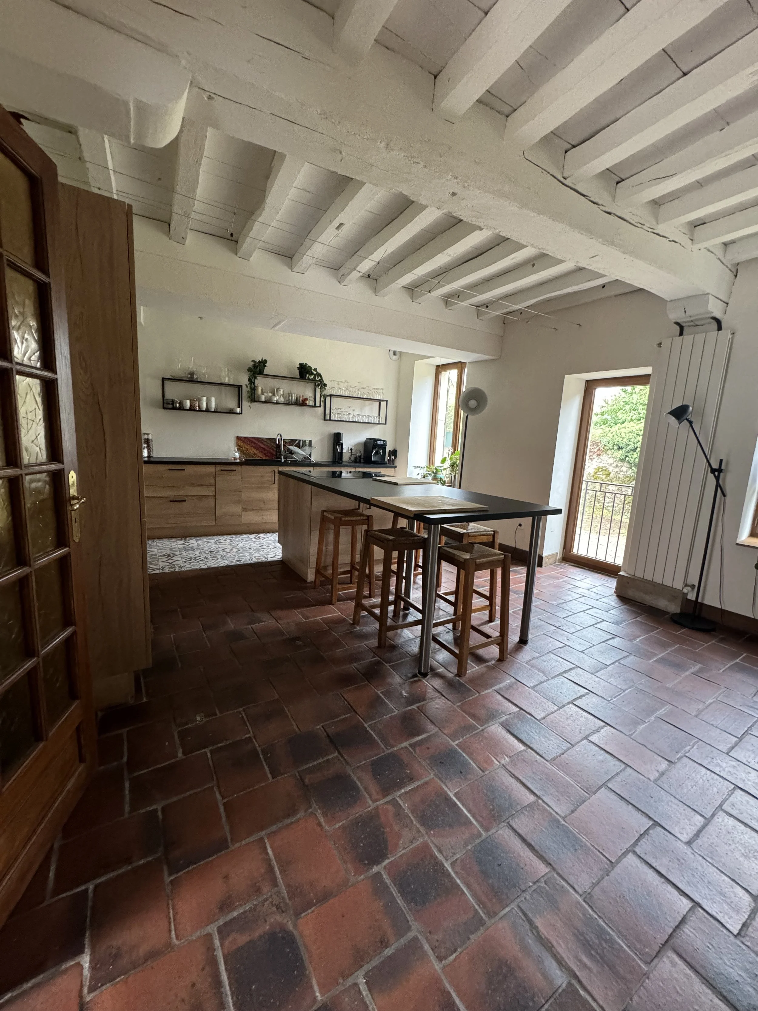 Cuisine rustique avec plafond en bois blanc, sol en carreaux de terre cuite, comptoir en bois, et deux fenêtres laissant entrer la lumière naturelle.
