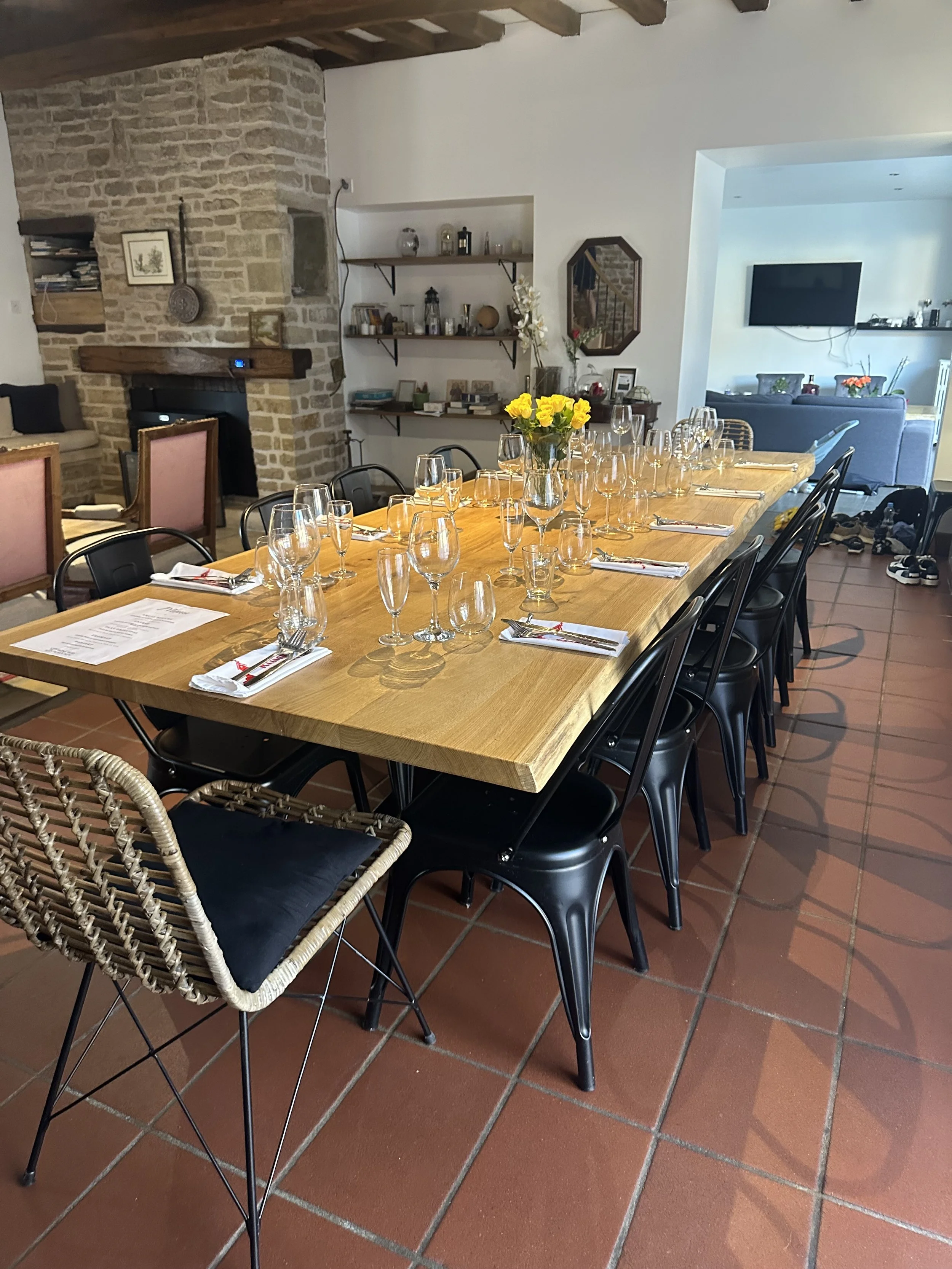Table long en bois avec verres à vin, bouteilles d'eau, fleurs jaunes en vase, couverts et napperons blancs dans une salle à manger élégante.